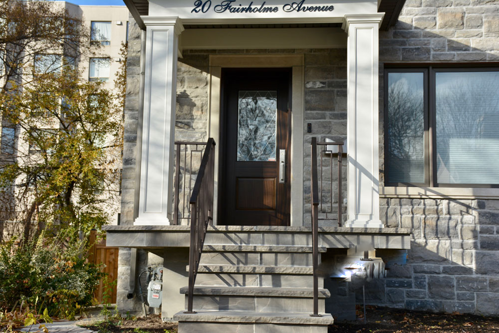 Stone house entrance with dark wooden door, white columns, and steps with metal railings. Address "20 Fairholme Avenue" is visible above the door. Large window on the right, trees and another building in background.