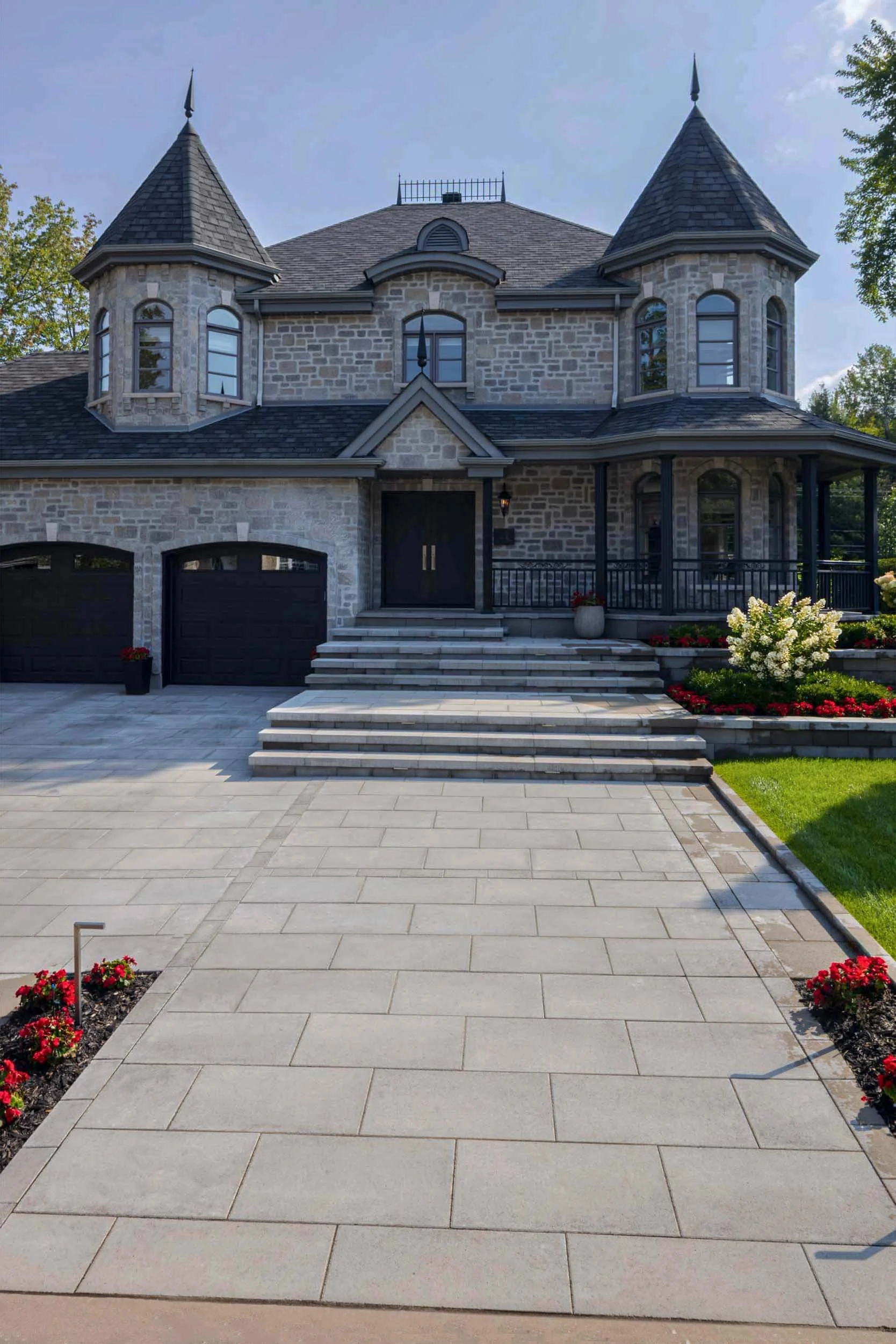 Two-story stone house with turrets, black double garage doors, and a large paved driveway lined with flowers.