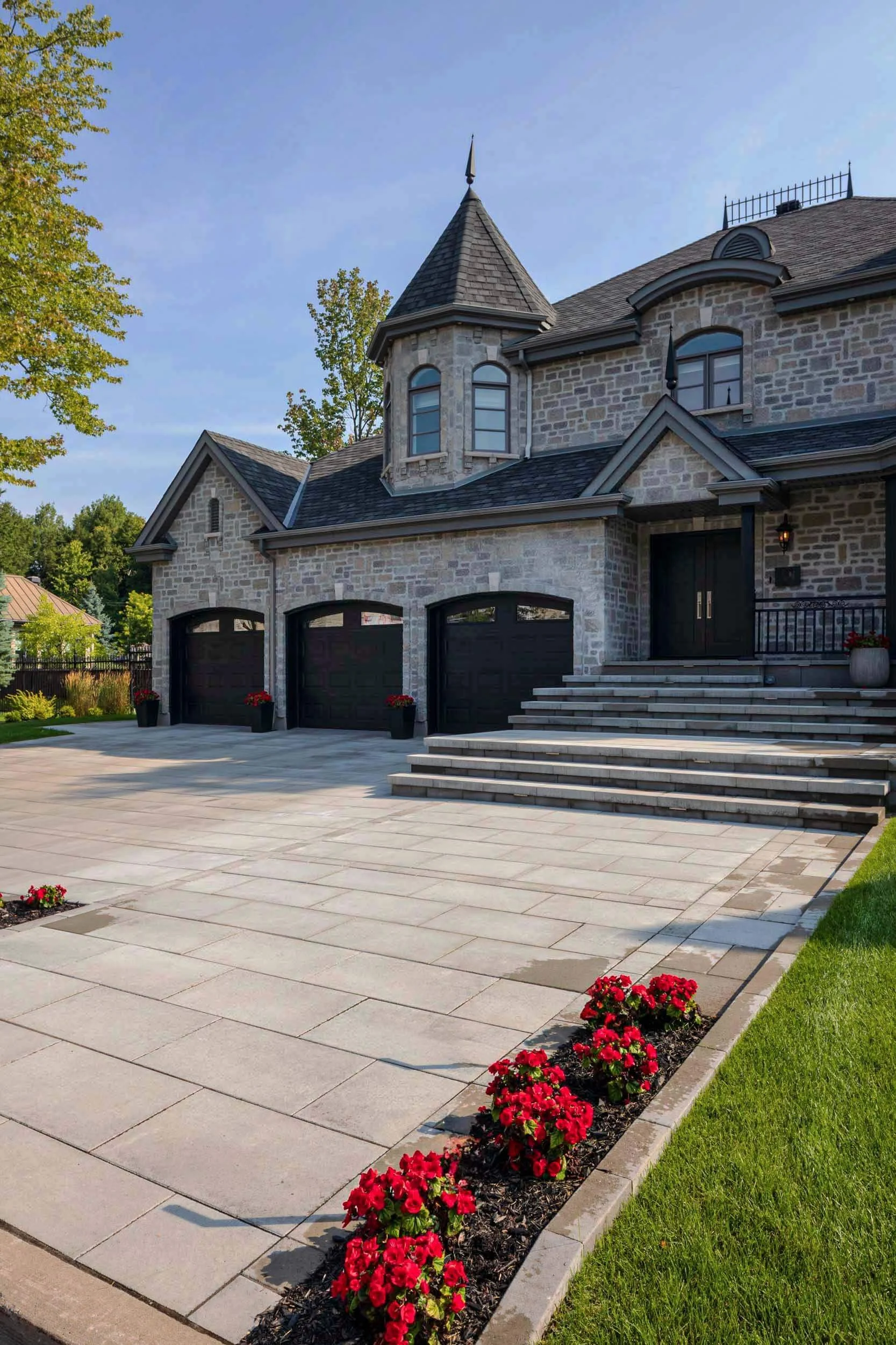 Stone house with a turret, three black garage doors, and a large driveway. Red flowers line the driveway's edge.