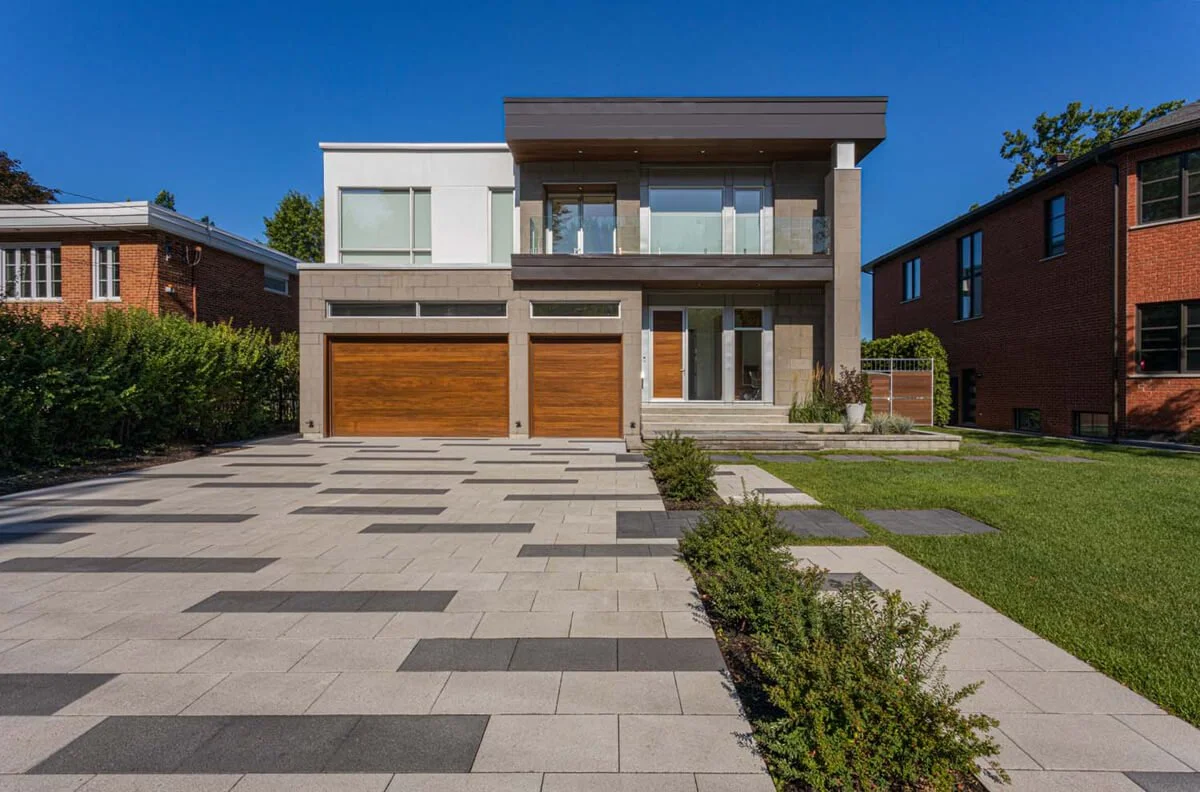 Modern two-story house with large windows, wooden garage doors, and a paved driveway lined with shrubs, situated between two brick buildings, under a clear blue sky.