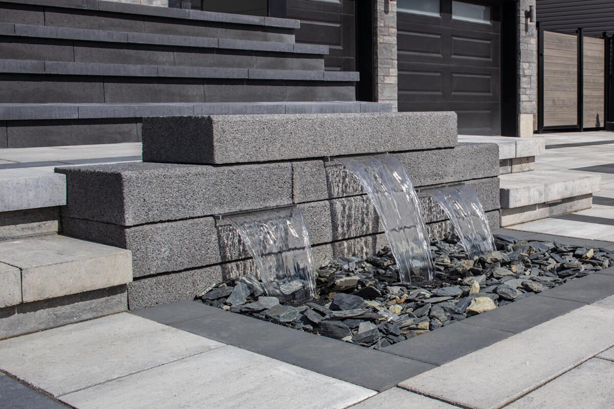 Modern stone water feature with three cascading streams over gray rectangular blocks into a bed of dark slate rocks, set in a concrete and stone patio.