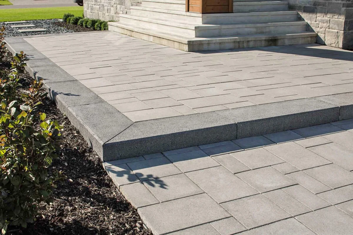 Gray stone patio with steps leading to a wooden door, surrounded by landscaping with shrubs and mulch.