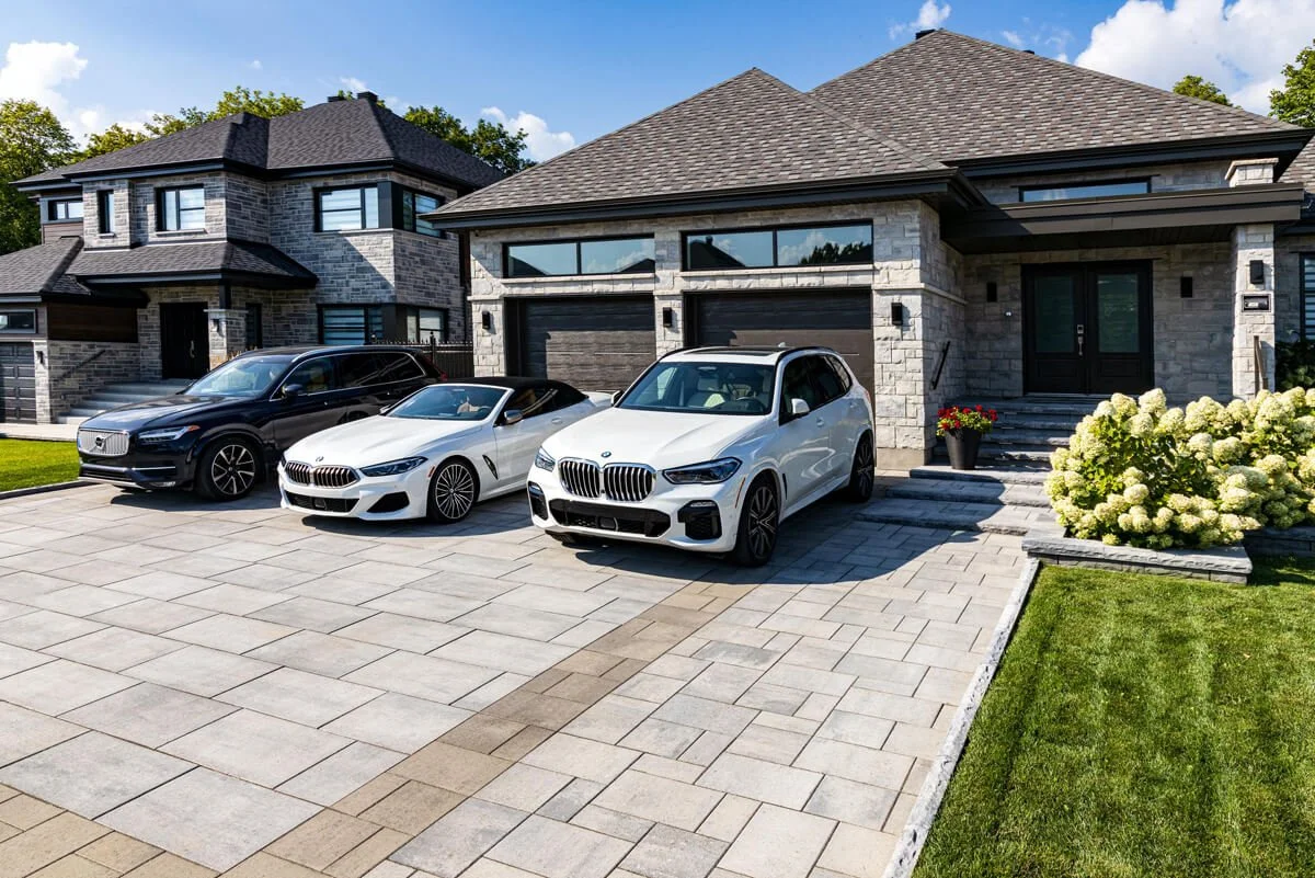 Luxury cars parked in front of two modern stone houses with a landscaped driveway and green lawn.