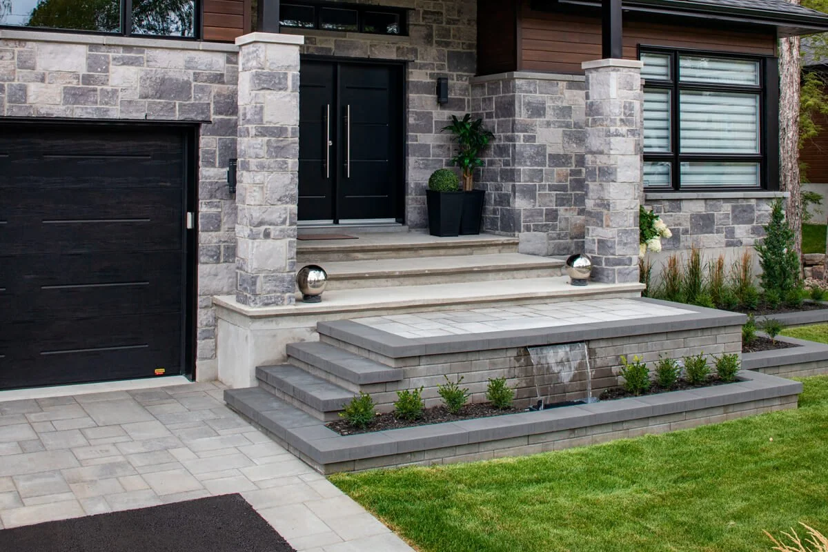 Modern house entrance with stone facade, black garage door, and double front doors. Steps lead to the entrance with decorative stone spheres. A small water feature and well-maintained greenery enhance the landscaping.