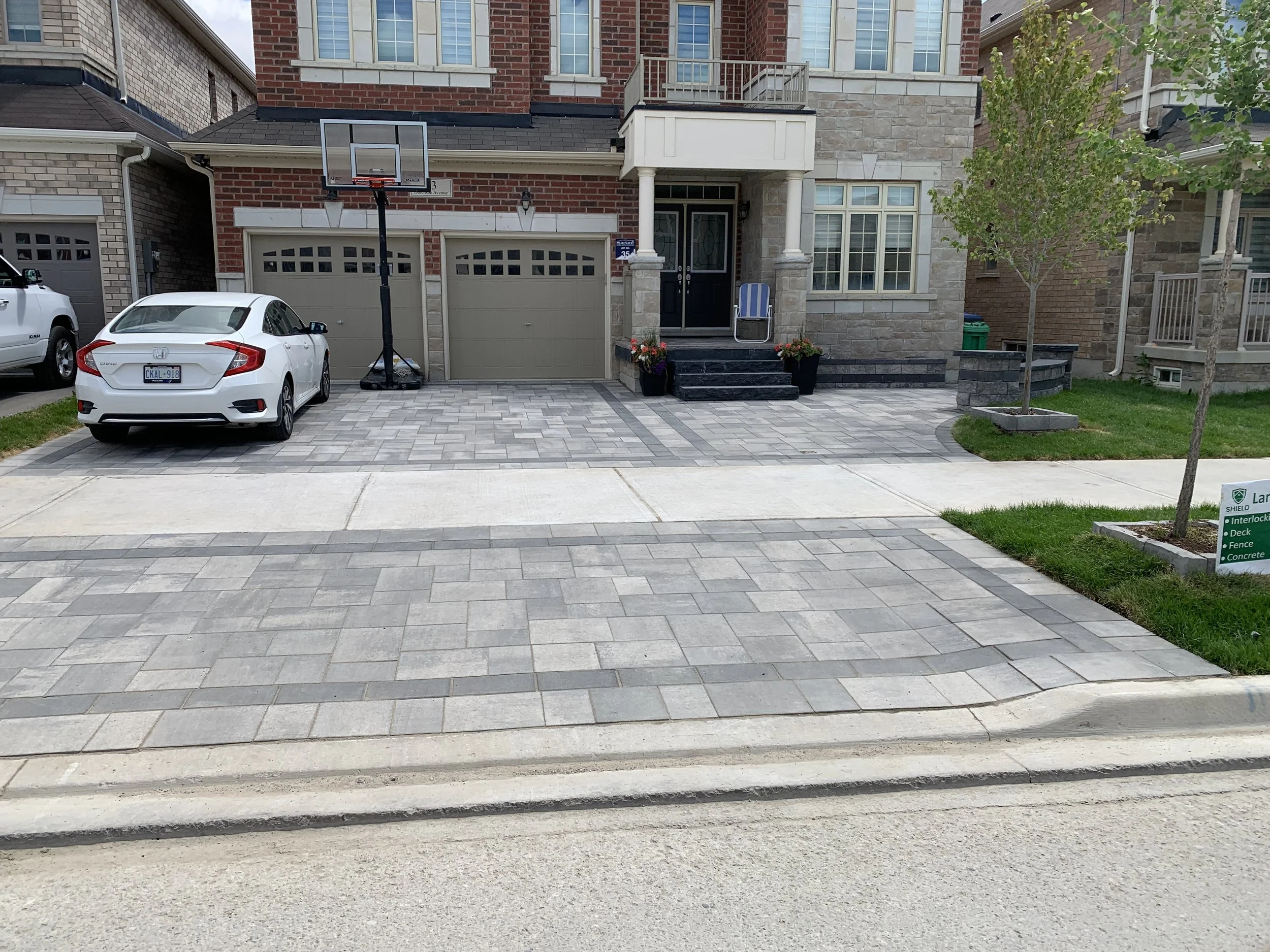 A suburban house with a brick and stone exterior, featuring a large driveway paved with interlocking stones. A white car is parked in front of a basketball hoop. The entrance has steps with potted plants on either side.