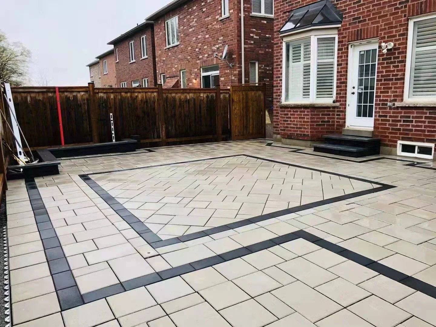 Backyard patio with light-colored paving stones, dark border lines, brick house, wooden fence, and steps to white door.