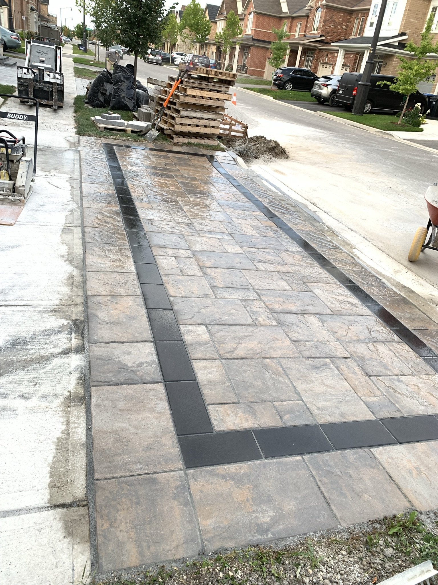 Freshly laid patterned paving stones on a driveway with construction materials nearby, including pallets and tools, in a suburban neighborhood.