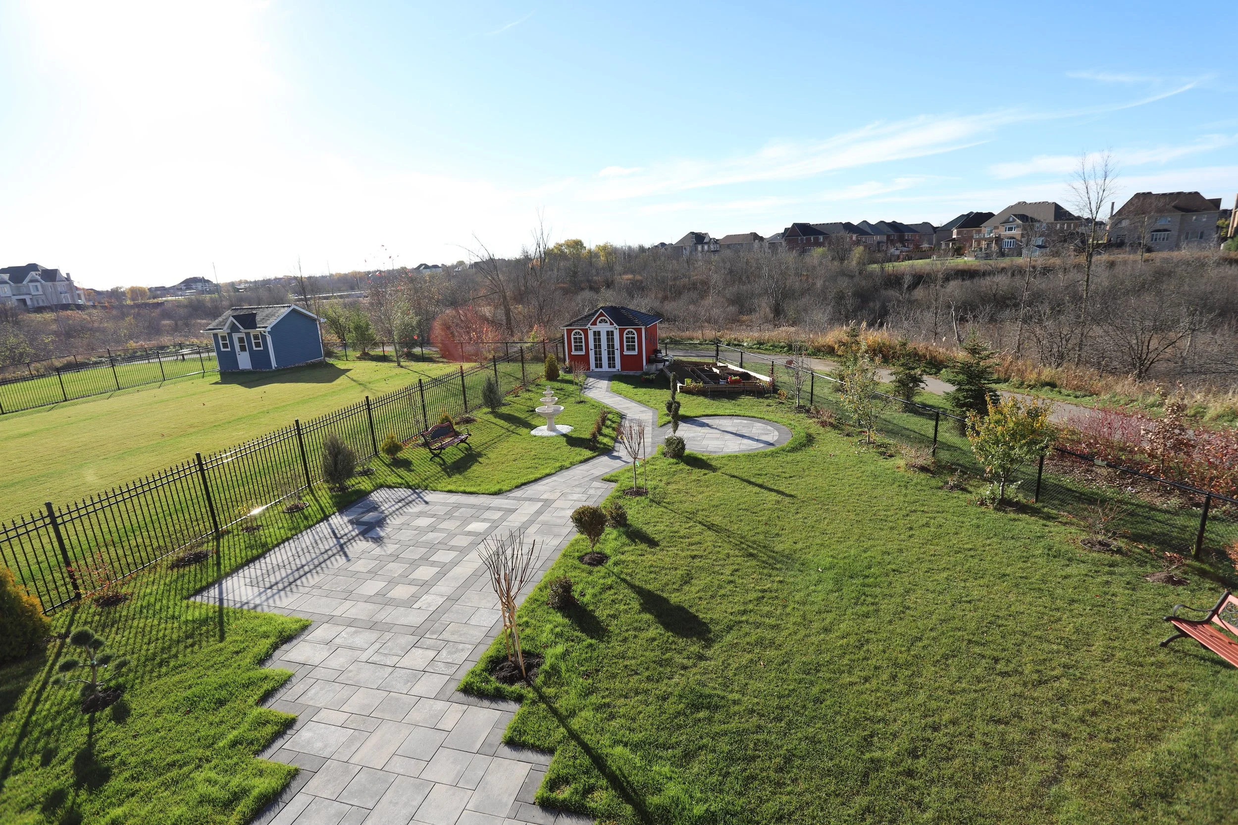 Spacious backyard with grass and paved path, red garden shed, blue shed, benches, and decorative fountain.