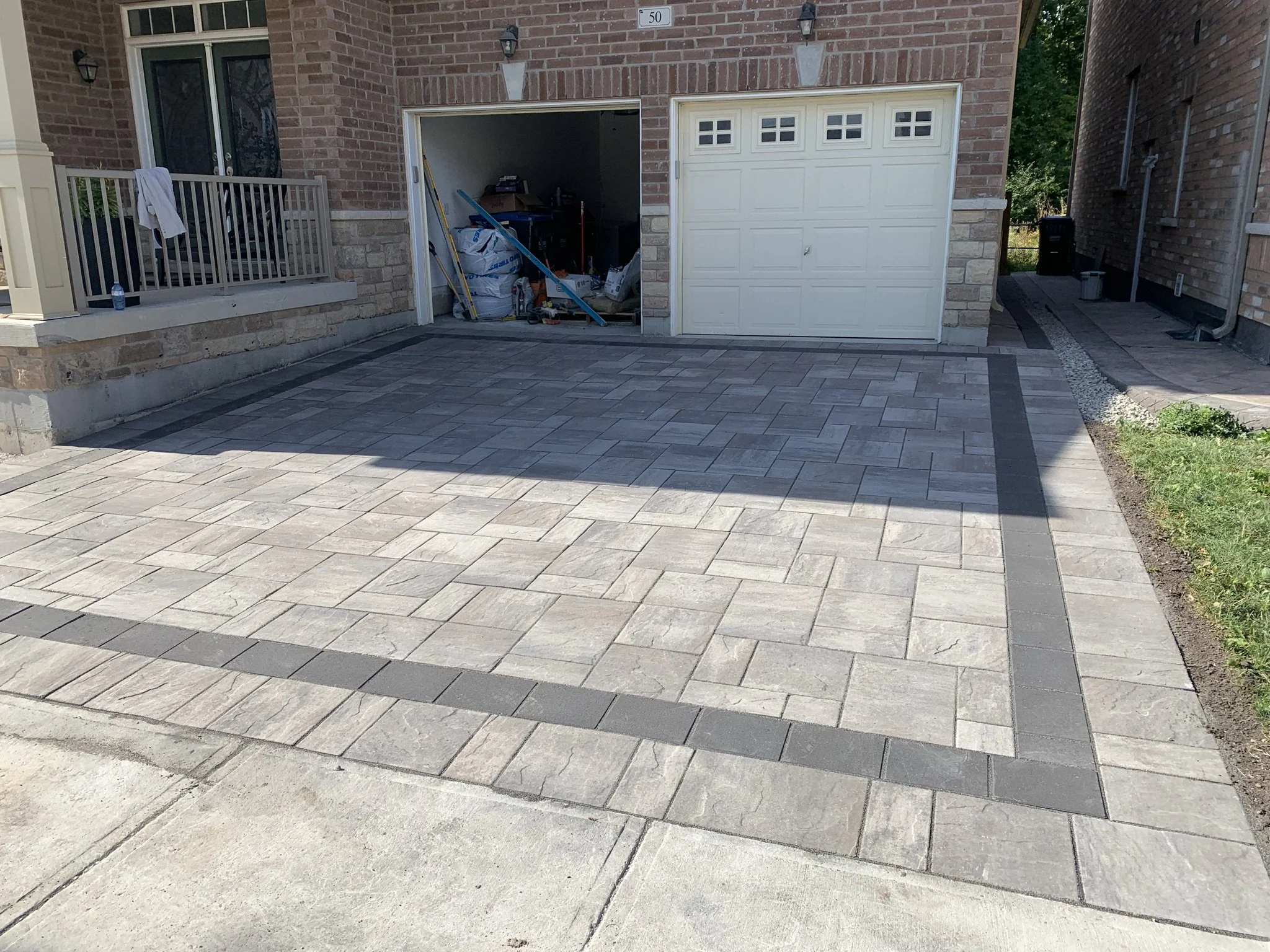 A newly paved driveway with interlocking gray bricks bordered by darker bricks, leading to an open garage attached to a brick house with a small porch.