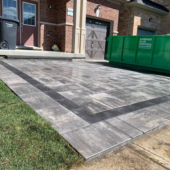 Newly installed gray tiled driveway with dark border, grass edge, red brick house, green waste disposal bin, trash can by entrance, and garage doors in view.
