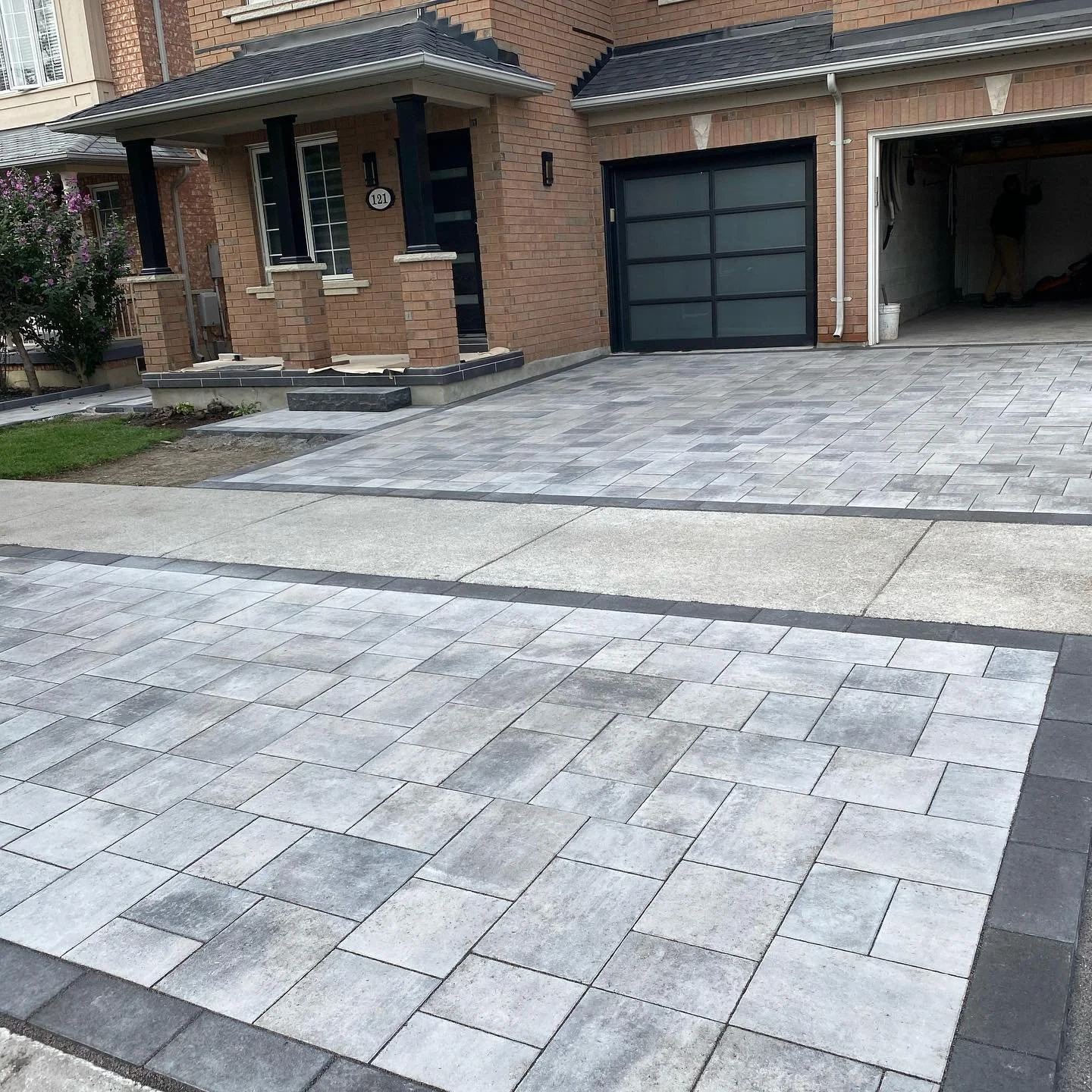 House with a paved driveway and garage, featuring gray interlocking stone tiles and a brick exterior.