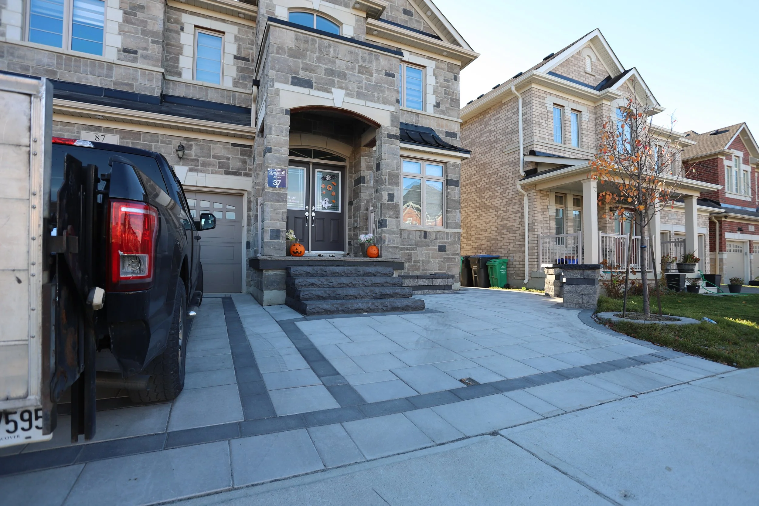 Brick house with a decorated Halloween porch, pumpkins, and a tree in front. Driveway with black vehicle parked.