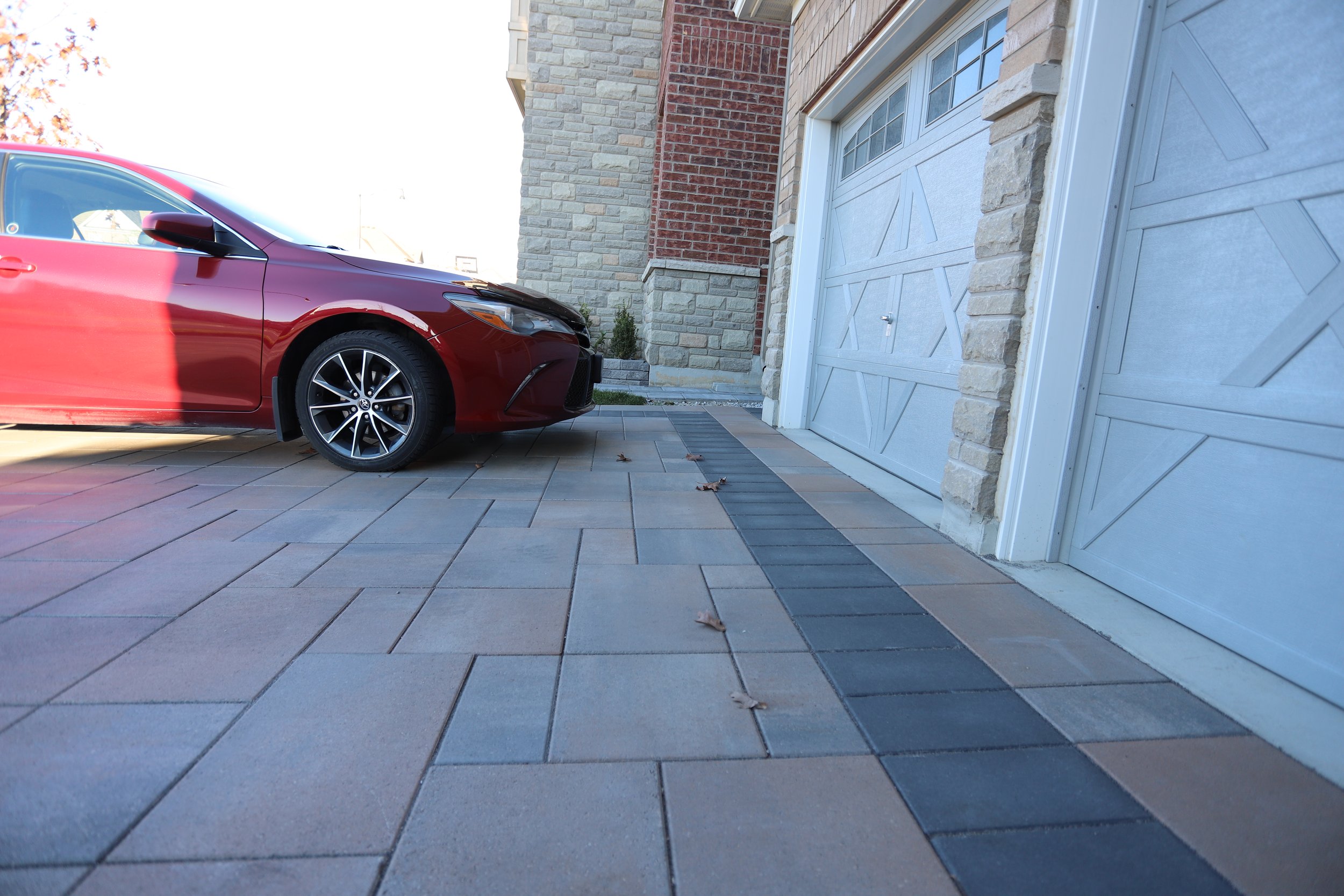 Red car parked in driveway next to a white garage door on a paved surface with stone and brick walls.