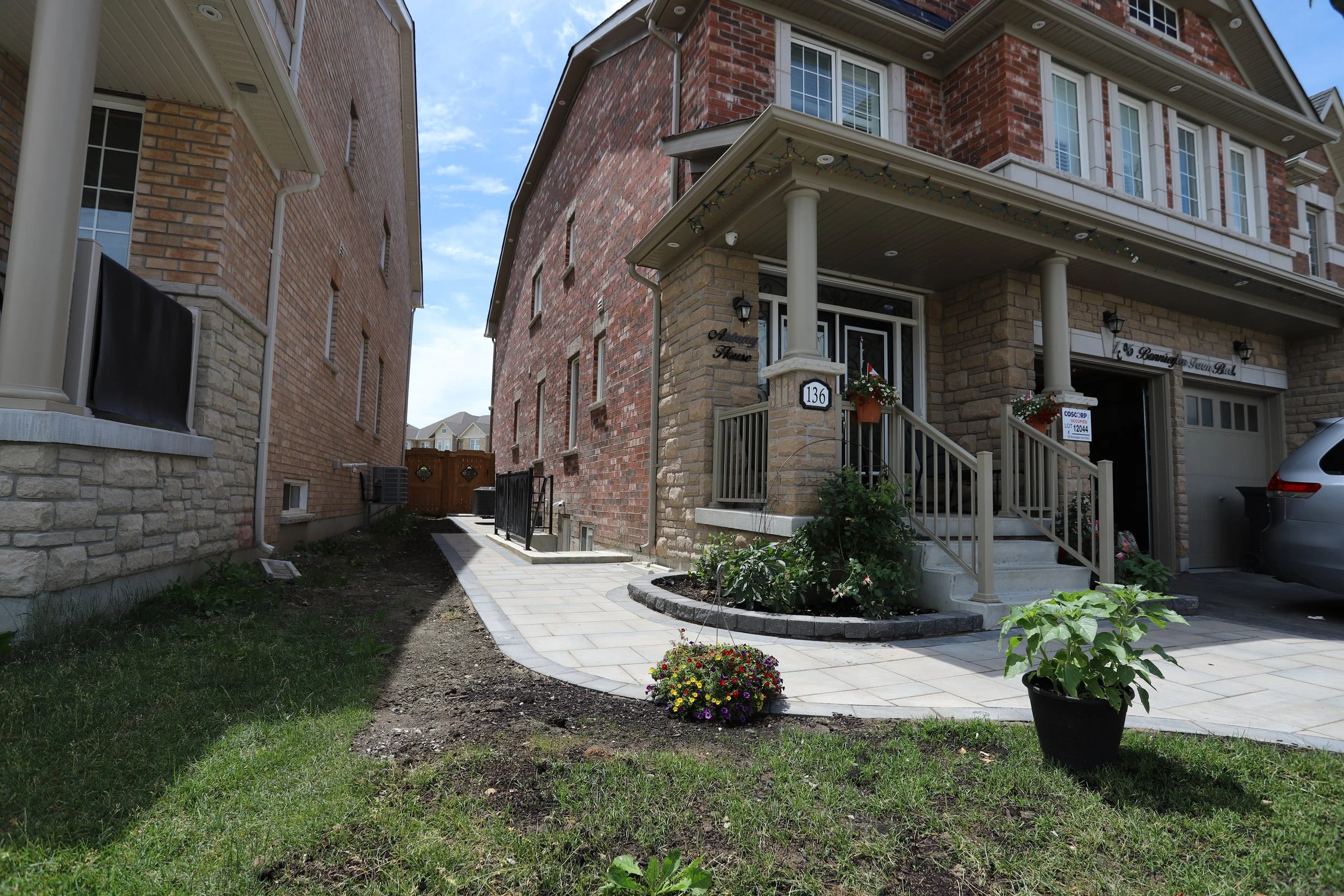 Suburban brick houses with front porches, a driveway with a car, potted plants, and a gate in the background.