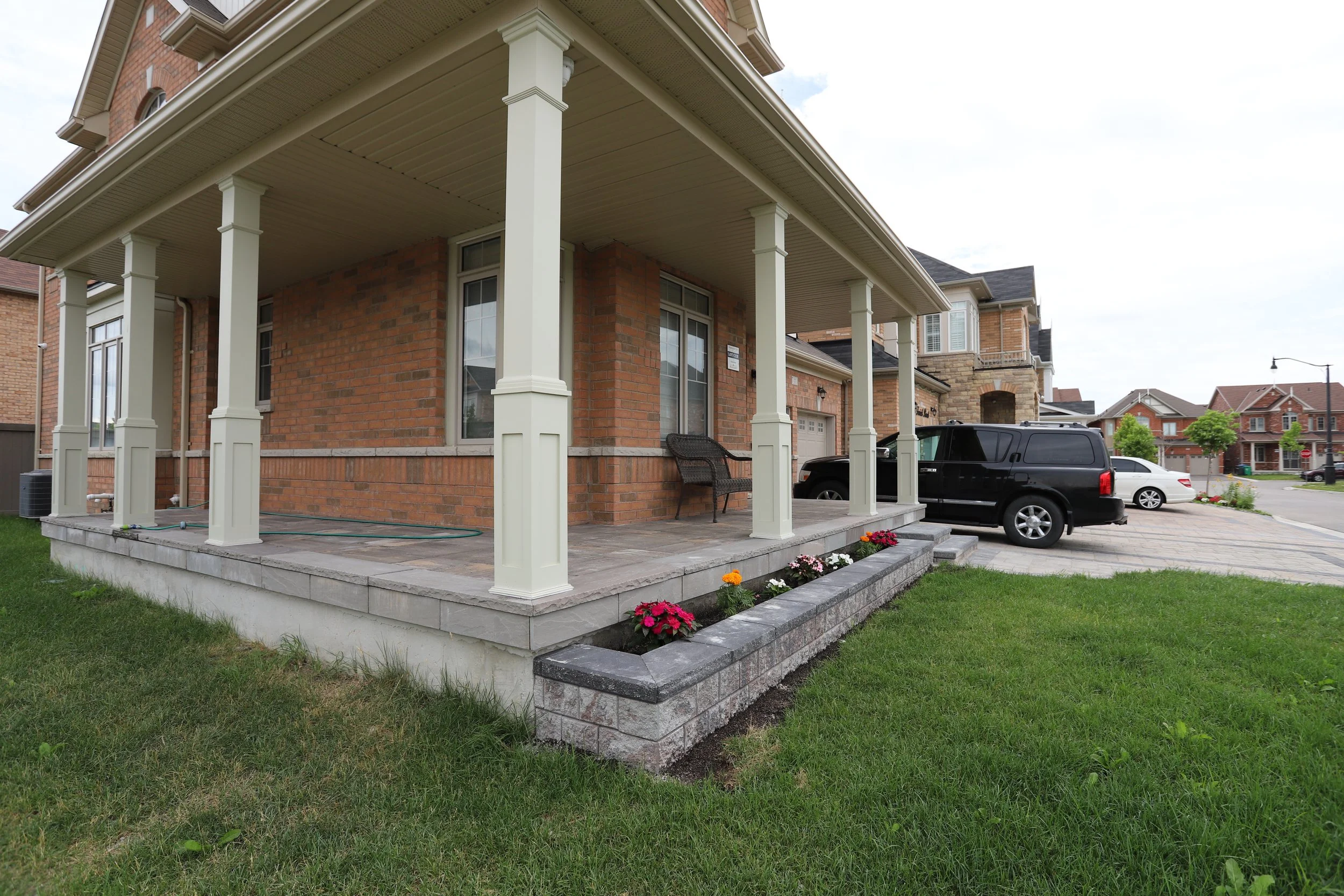 Suburban house with covered porch, brick exterior, and flower bed; black SUV and white car in driveway.
