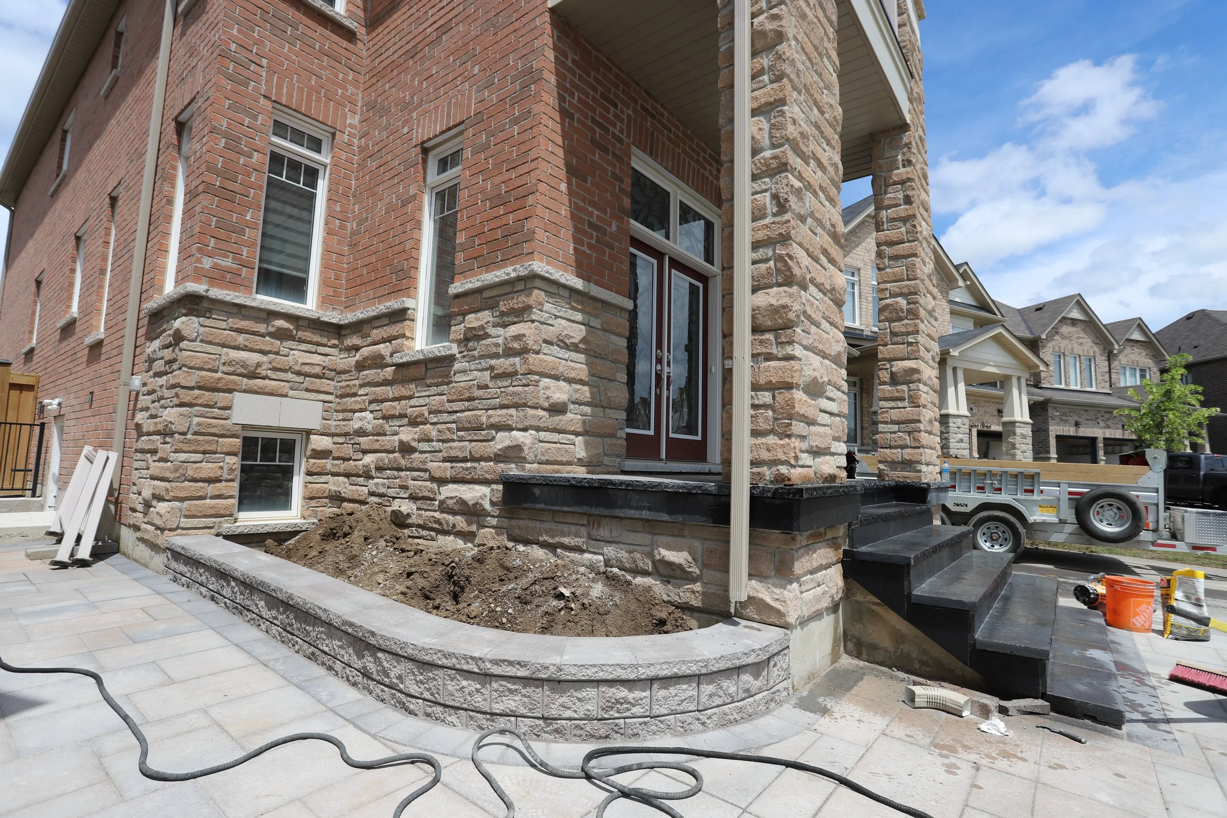 House exterior under renovation with stone and brick facade, black steps, and construction materials including a trailer, hose, and tools.
