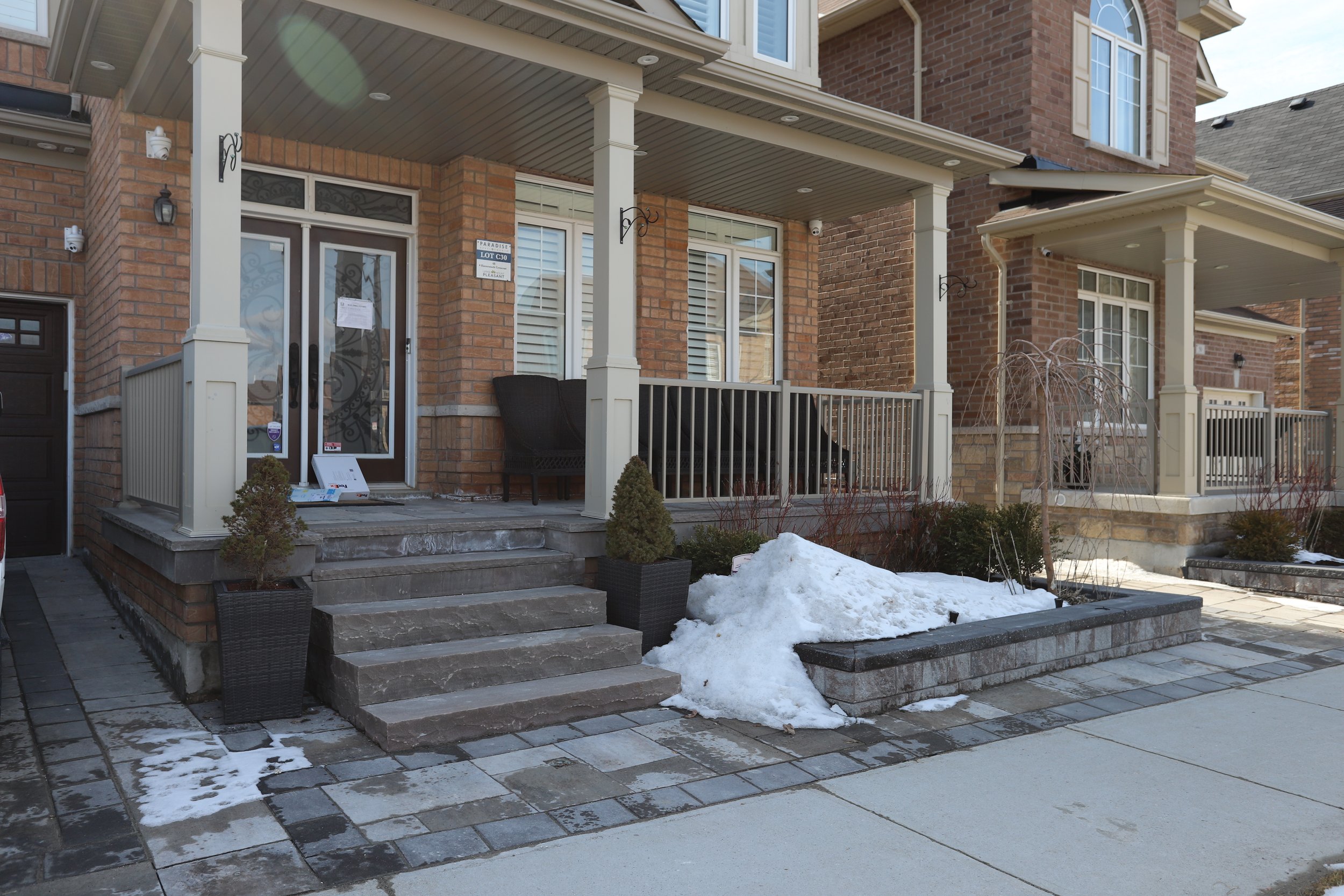 Front porch of a suburban house with brick exterior, snow on the walkway, potted plants, and a black chair.