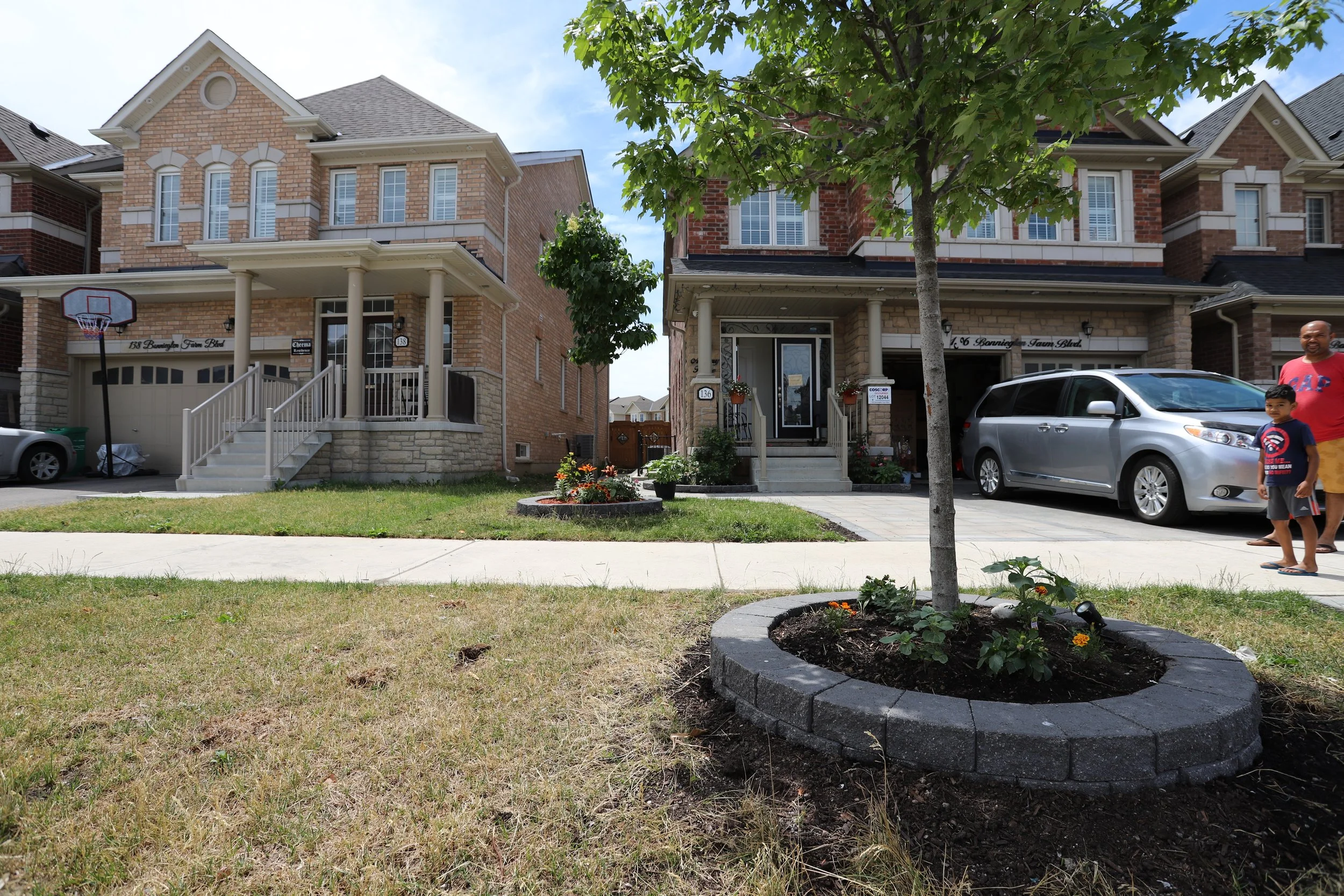 Two suburban houses with brick facades, a tree in a front yard surrounded by flowers, and a man with a child near a parked silver minivan.