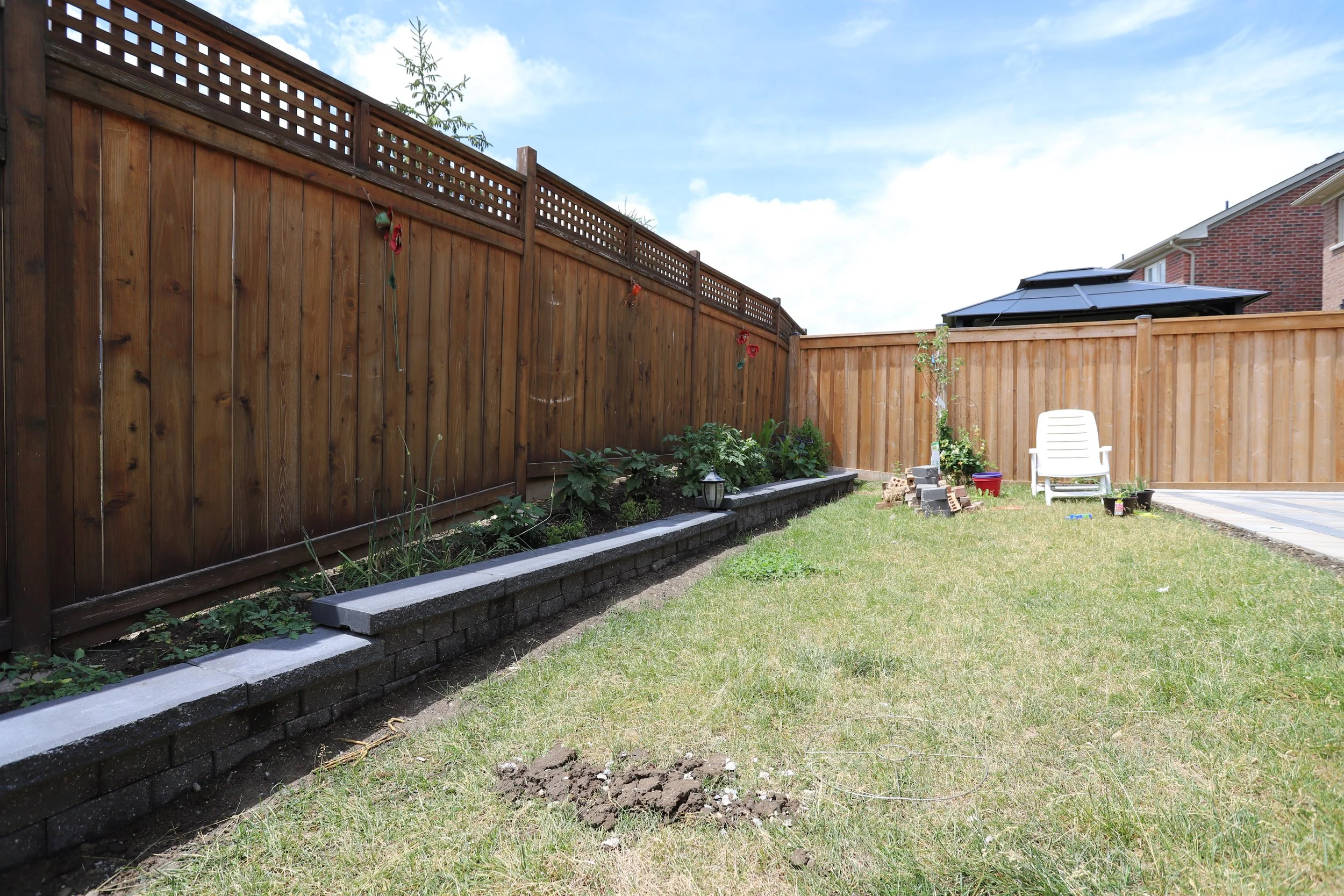 Backyard with wooden fence, grassy lawn, flowerbed, white plastic chair, and patio area under a blue sky.