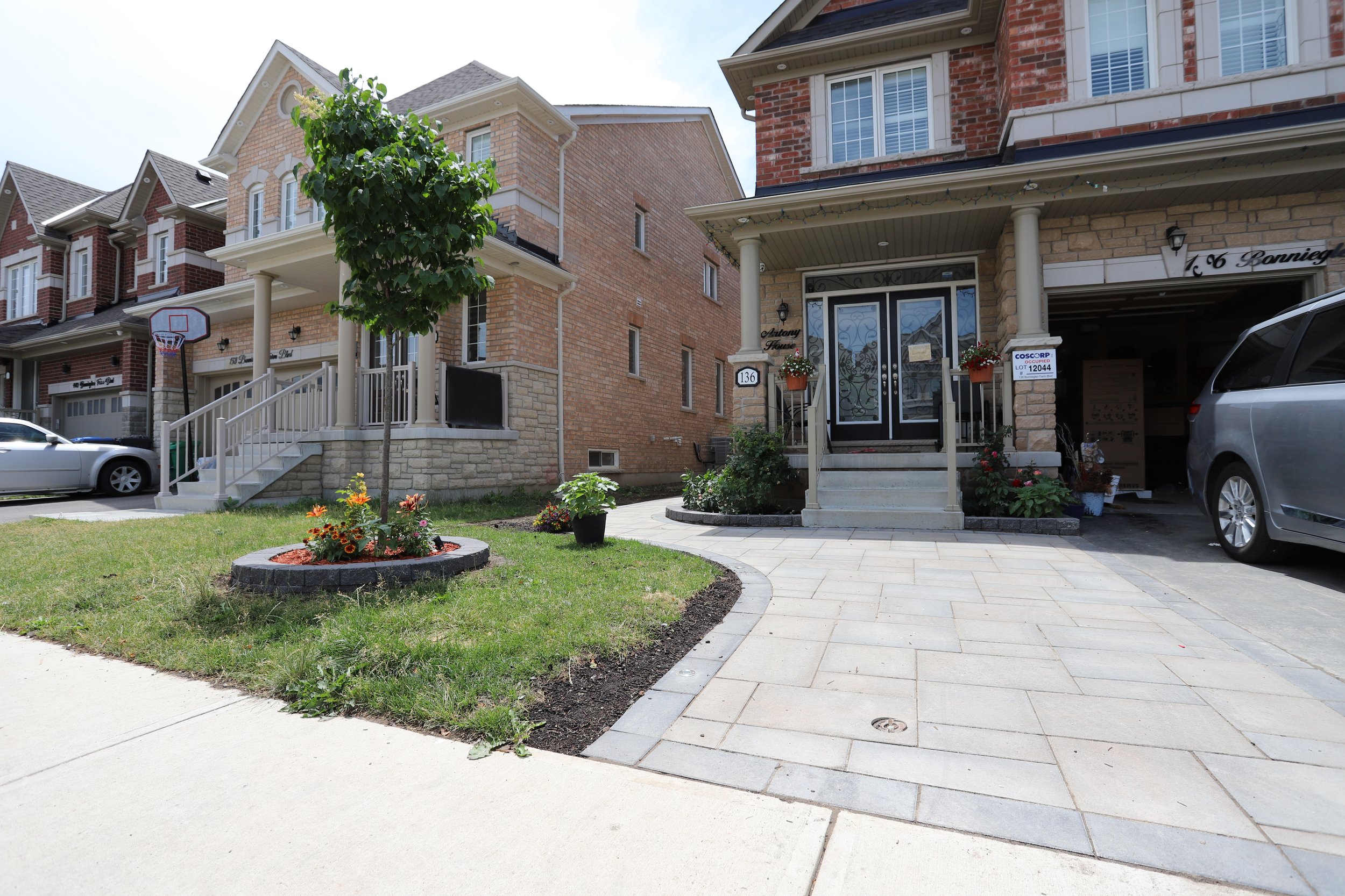 Suburban neighborhood with three brick houses, landscaped lawns, and a driveway with a parked van. A basketball hoop is visible in one driveway, and a tree with flowers is planted in front of the nearest house.