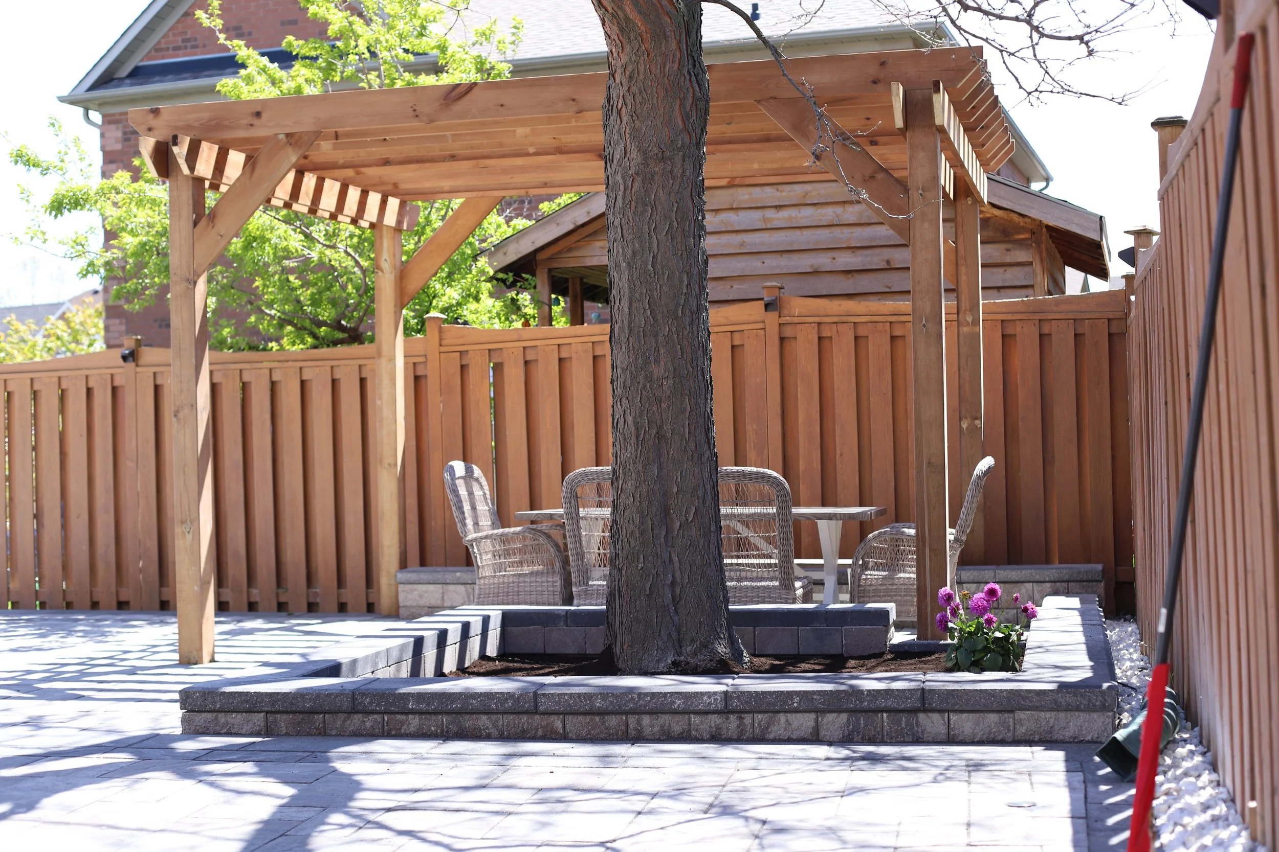 Backyard patio with wooden pergola, tree, wicker chairs, stone flooring, and flower bed.
