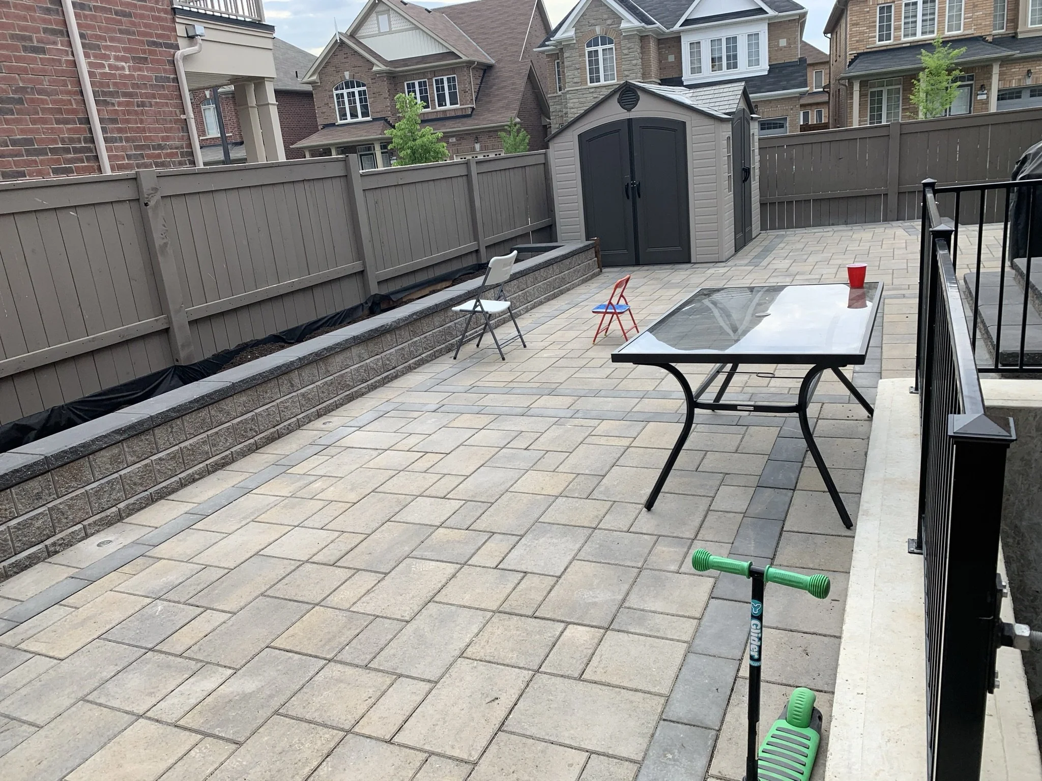 Backyard patio with a paver stone surface, glass table, folding chairs, a scooter, and a storage shed against a fenced backdrop of residential houses.