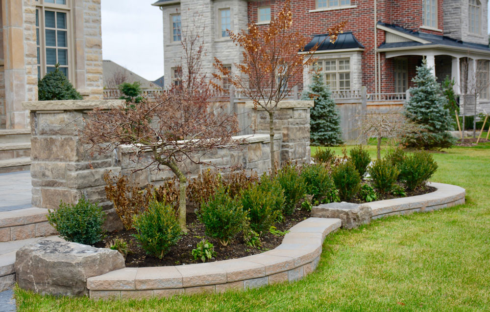 A landscaped front yard with trimmed bushes, small trees, and a stone patio. The yard has a manicured lawn and is bordered by stone walls. Two houses with brick and stone exteriors are in the background.