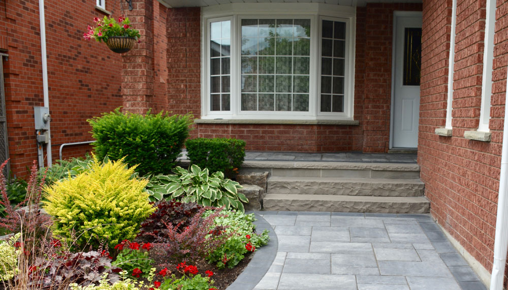 Front entrance of a red brick house with a paved walkway, steps, and landscaped garden featuring various shrubs and flowering plants.