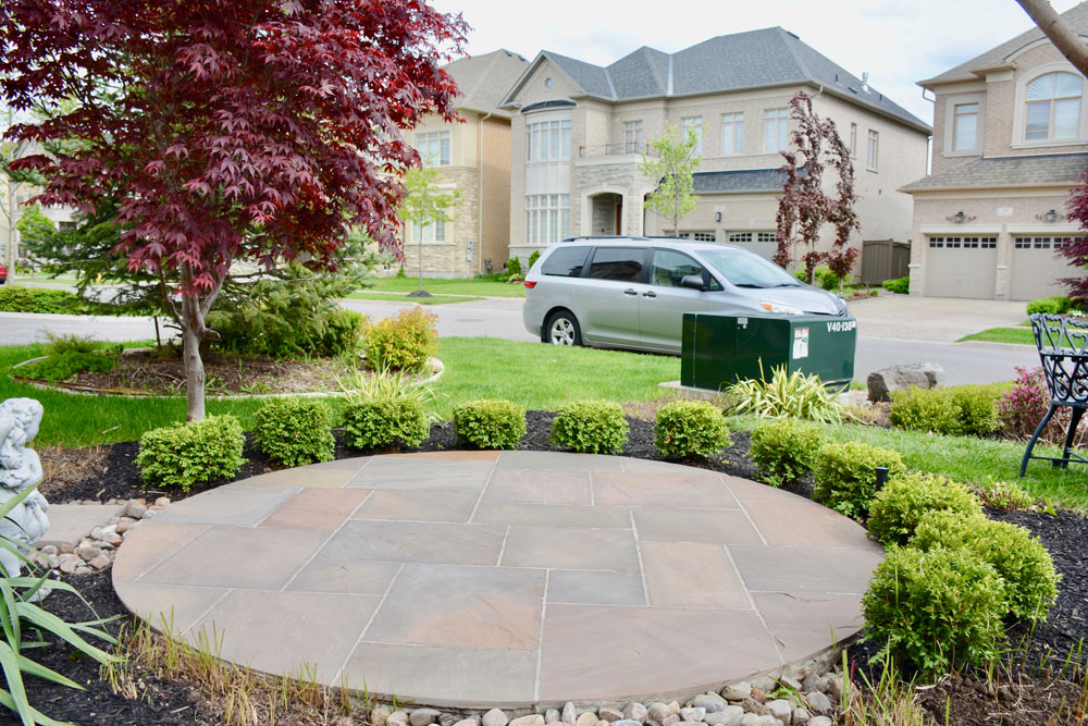 Circular stone patio with greenery, a red tree, suburban houses, and a parked van in the background.