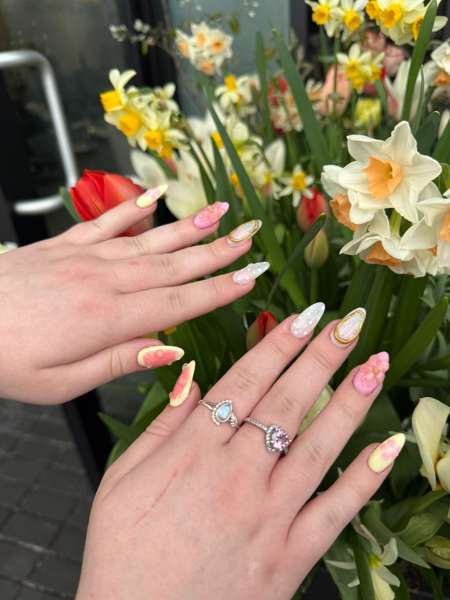 Two hands with decorated nails and rings placed over a bouquet of colorful spring flowers, including daffodils, tulips, and other flowers.