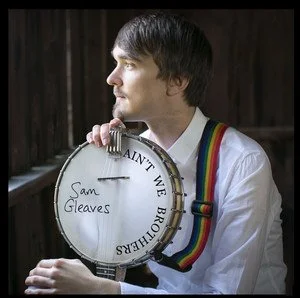 A man holding a tambourine with a handwritten message reading 'Saint We' and 'Sam Gleave's' in front of a window.
