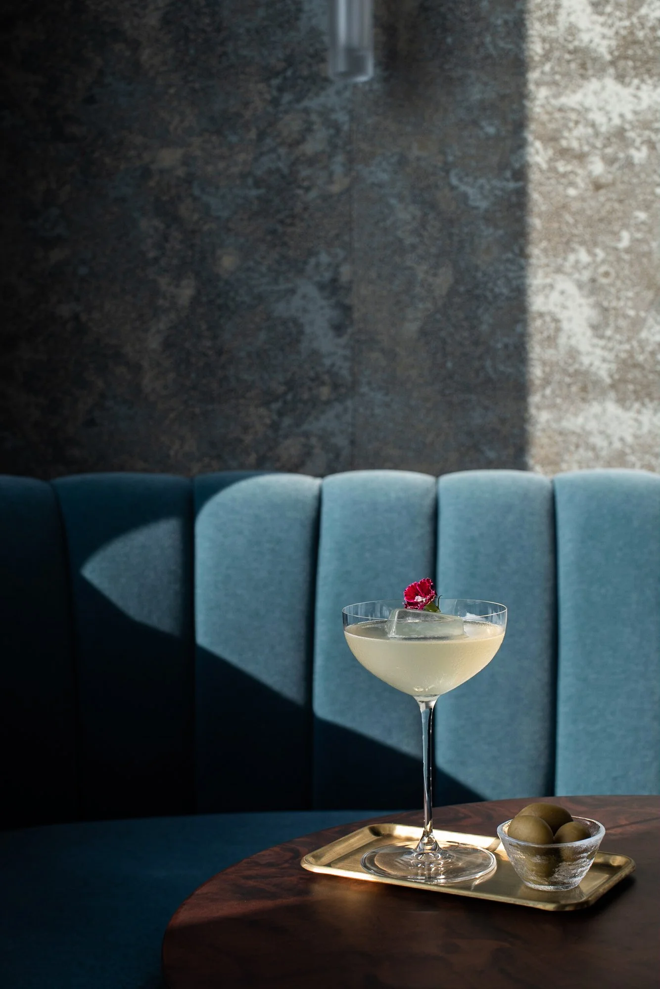 A cocktail in a coupe glass garnished with a pink flower on a tray, with a small bowl of green olives on the side, on a wooden table with a blue upholstered bench and a dark stone wall in the background.
