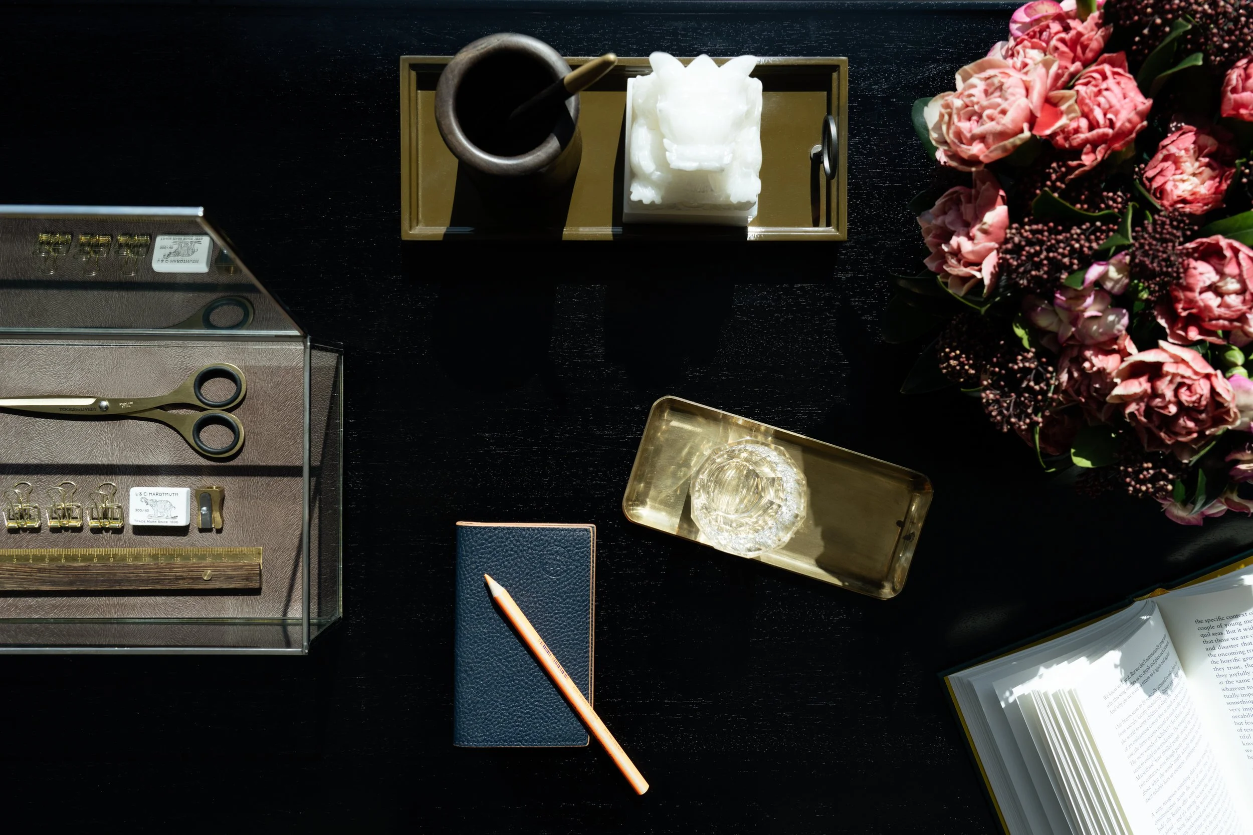 A black desk with various objects including pink flowers, a pair of scissors, a pen, a glass, a book, and decorative items.