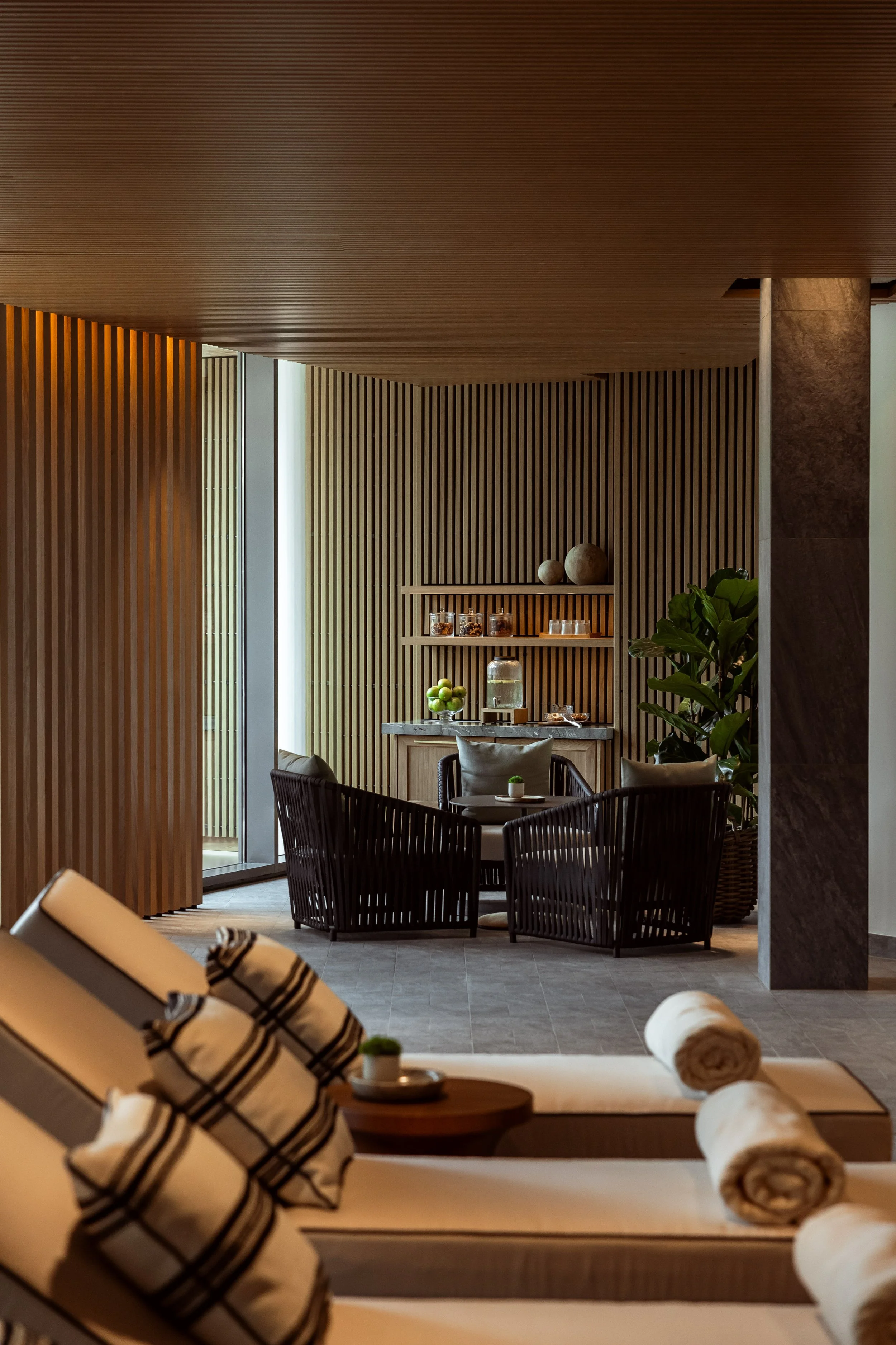 A modern, cozy lounge area with a wooden ceiling and walls, plush chairs, and decorative elements including plants, jars, and a bowl of green apples.