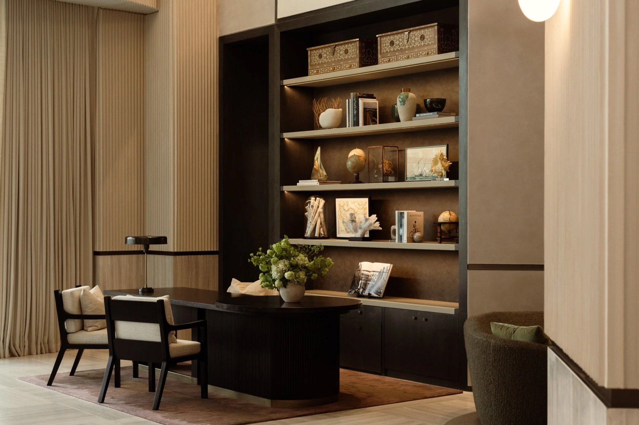 A modern living room with a round black dining table, white chairs, a large flower arrangement, and a dark wood bookshelf filled with decorative items and books. The room features beige and light wood tones with a textured wall and warm ambient light
