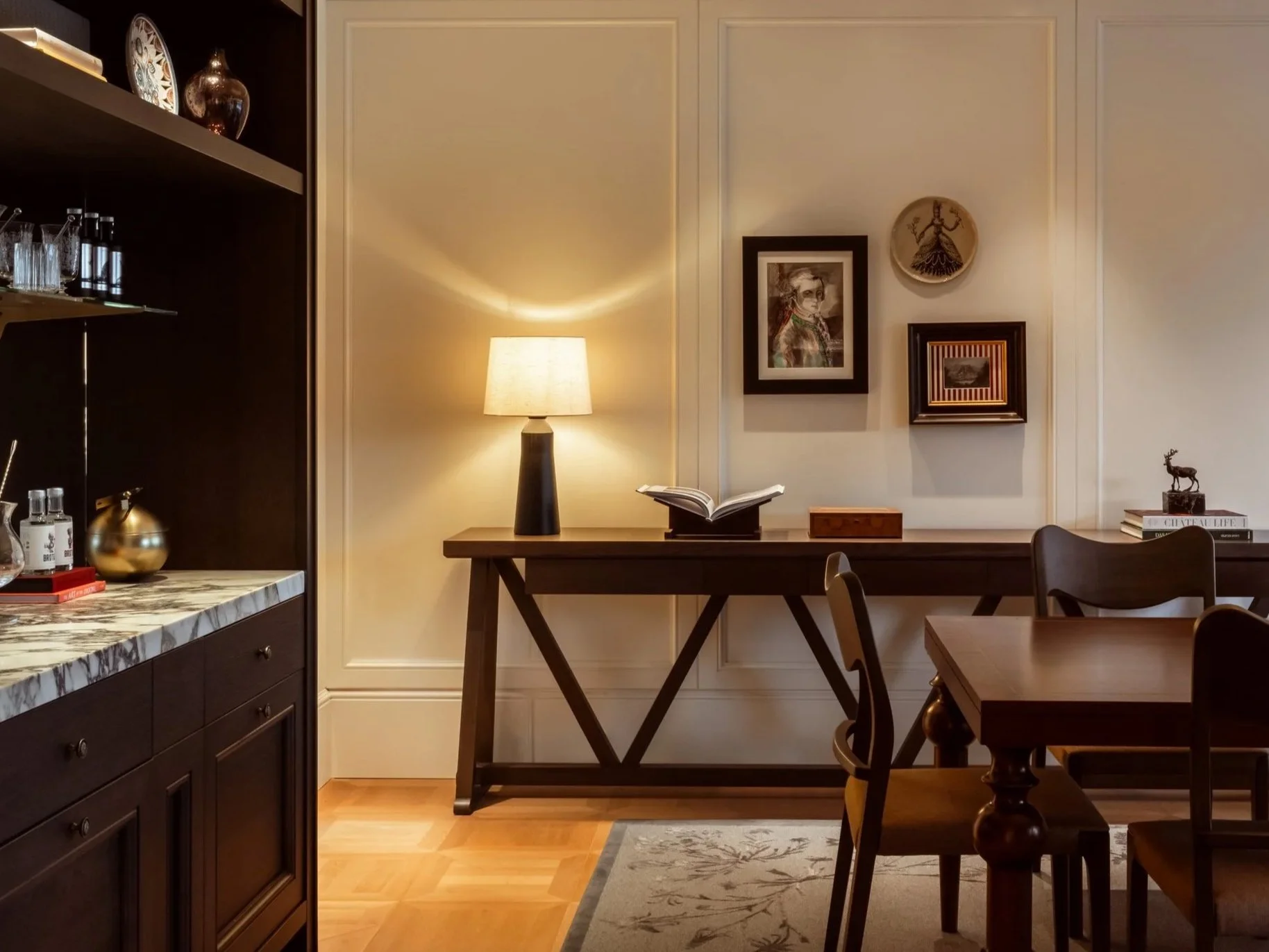 A cozy corner of a dining room showing a wooden table, chairs, a side table with a lamp, framed photos, and decorative wall art. A bookshelf with glassware and decorative items is visible on the left.