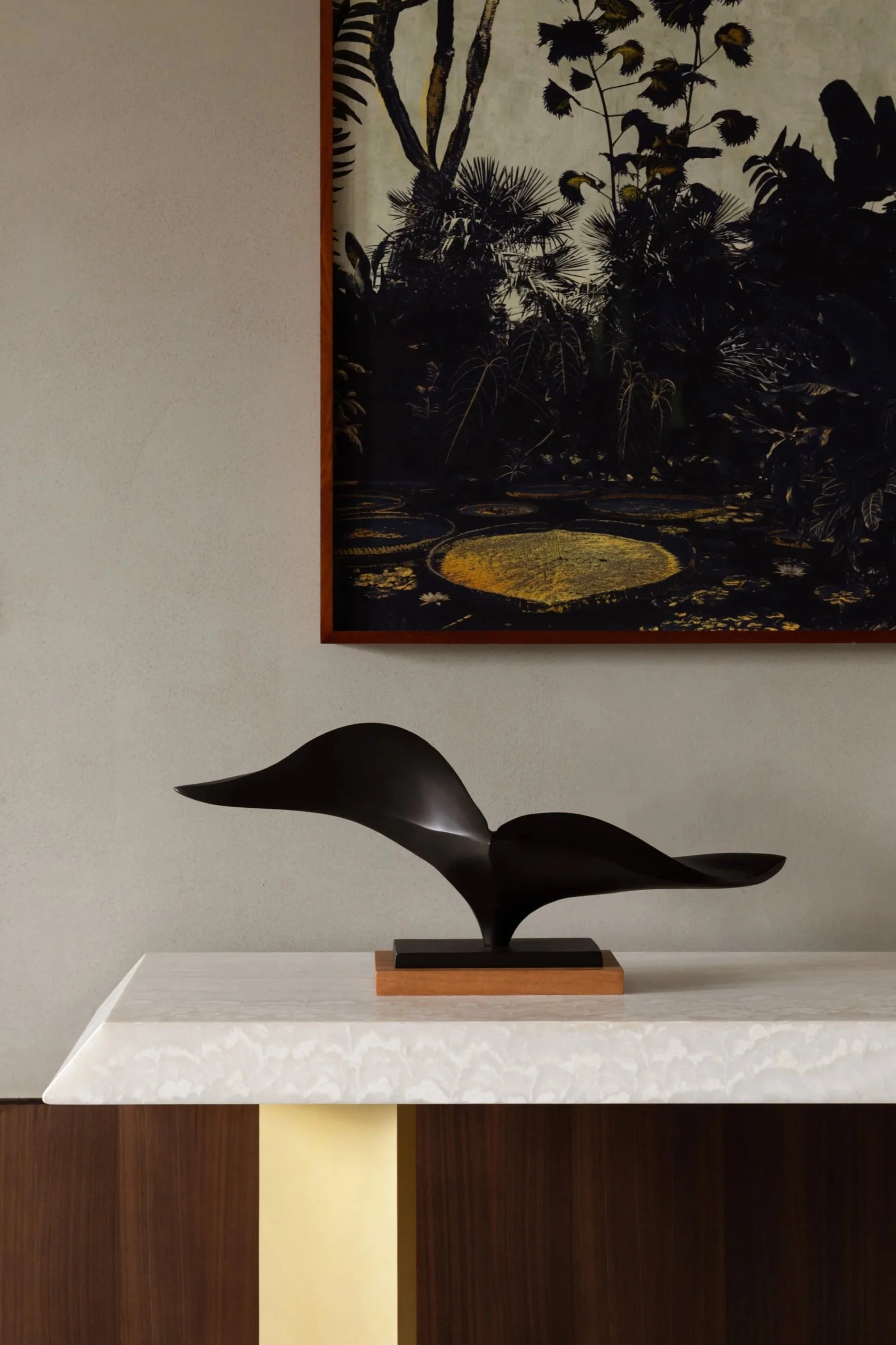A black, abstract sculpture resembling a whale with a white marble base, placed on a white table in front of a wall with a framed painting of a jungle scene with water lilies and lush tropical plants.
