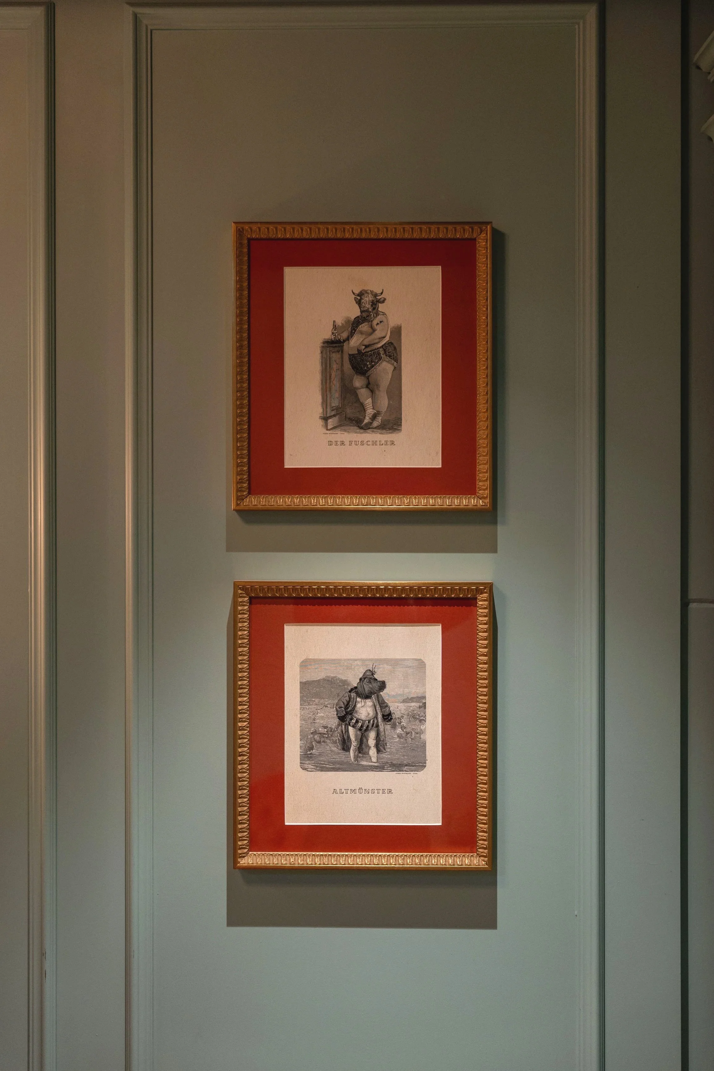Two framed artworks on a green wall. The upper artwork depicts a figure with a cow head, standing with crossed arms next to a wooden column, labeled 'DER FUSCHLER.' The lower artwork shows a bear in a landscape, labeled 'ALTMÜNSTER.'