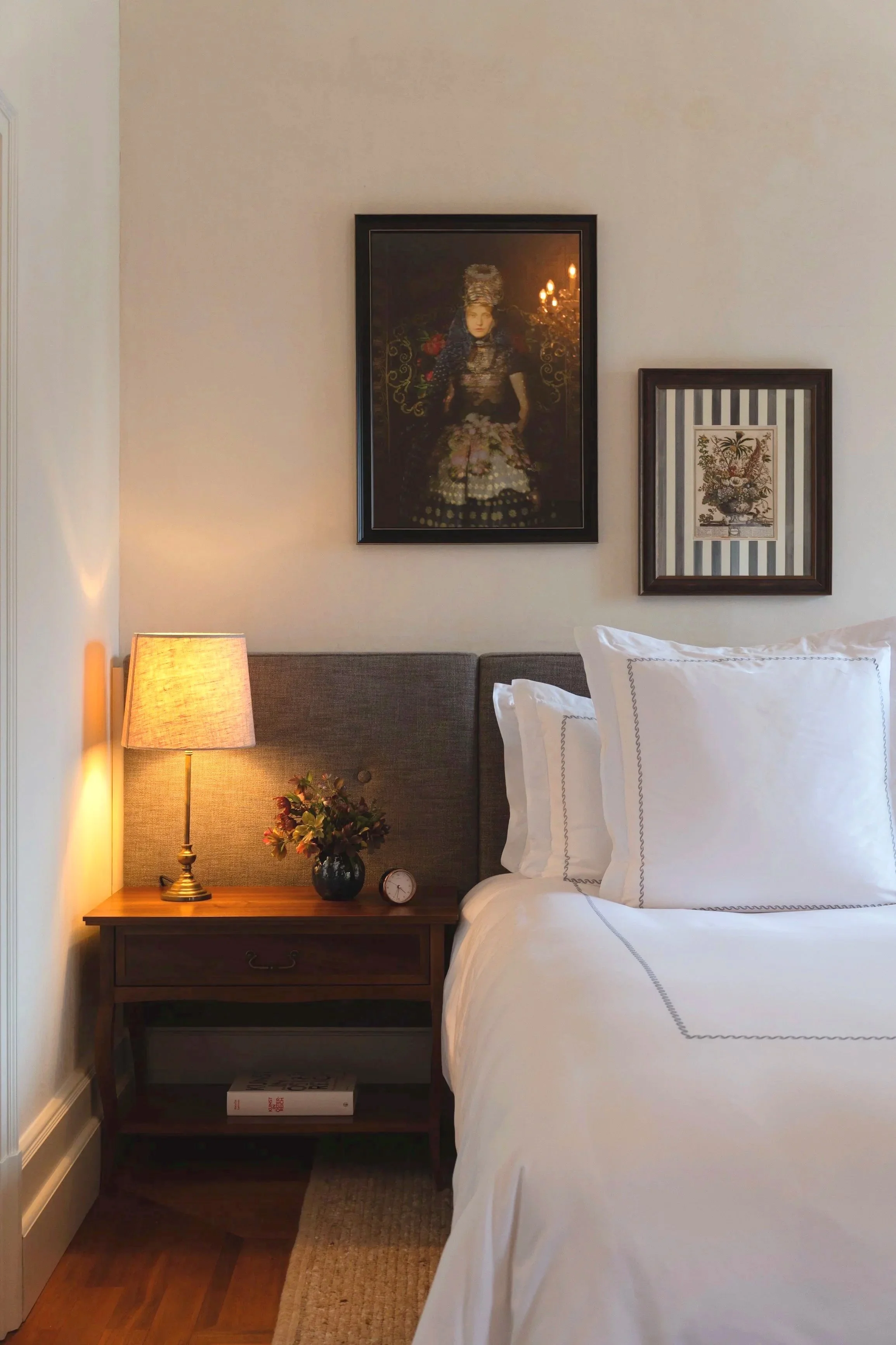 A cozy bedroom corner with a wooden nightstand, a beige table lamp, a vase with flowers, a small clock, a bed with white linens and pillows, two framed pictures on the wall, one of a woman in vintage clothing and the other with a floral design, and a