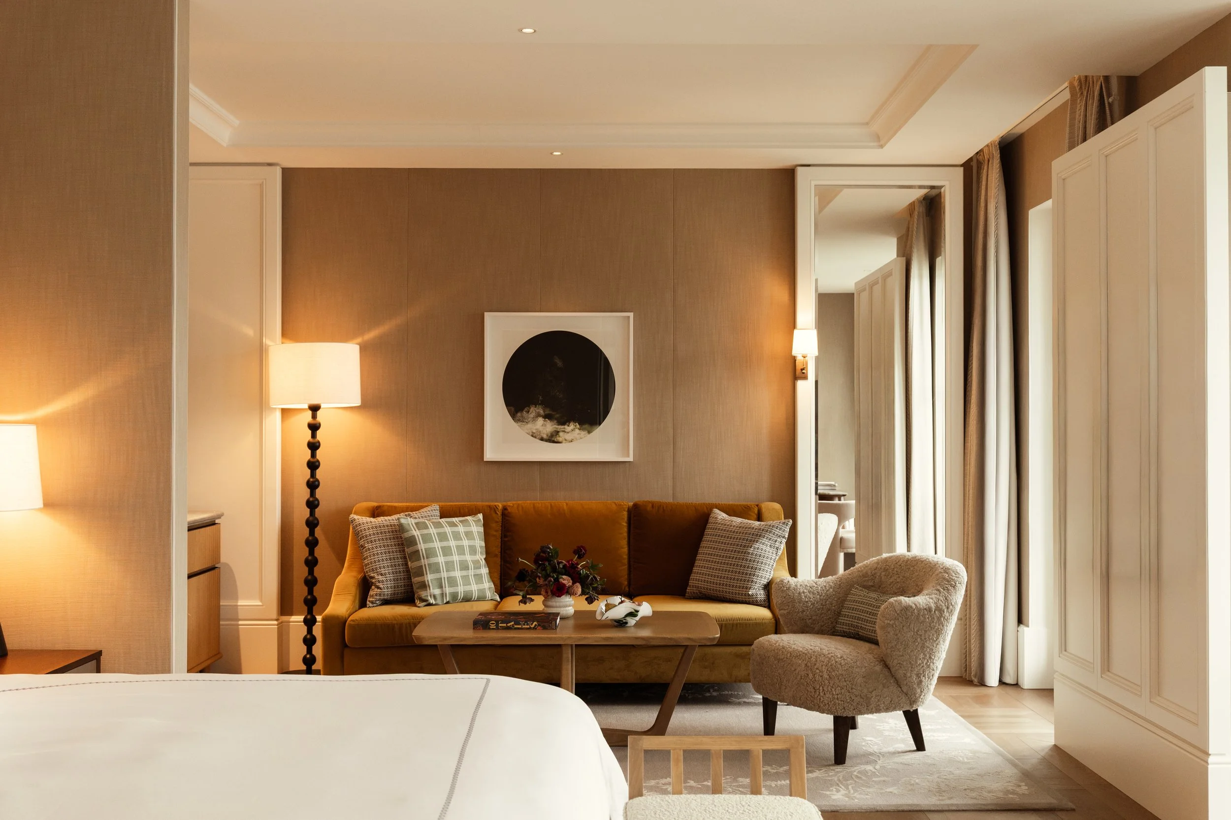 A cozy hotel room with a beige sofa decorated with plaid pillows, a beige armchair, a wooden coffee table, lamps, framed artwork, and large windows with beige curtains.