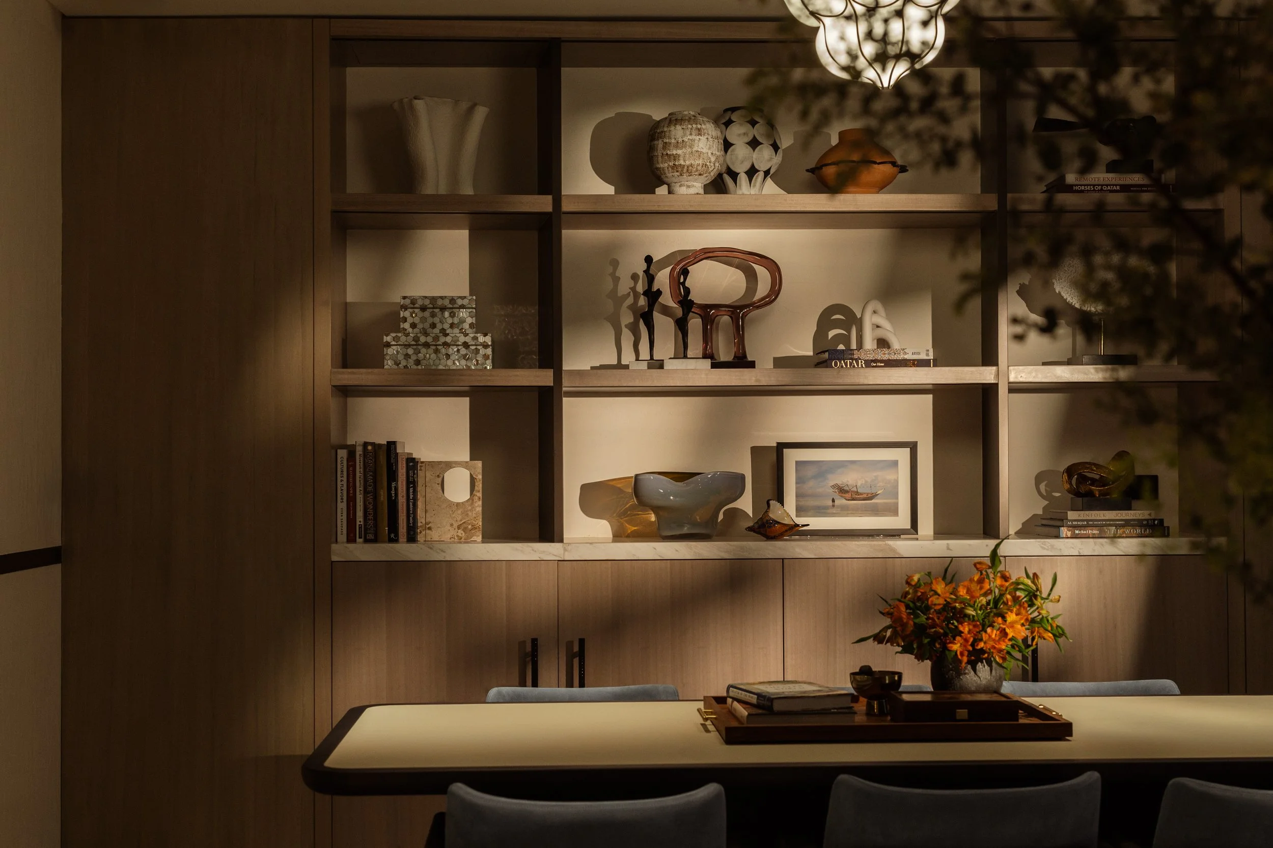 A living room or dining area with a wooden bookshelf filled with decorative vases, sculptures, and books. A table in the foreground has a tray with books or magazines, and a vase with orange flowers is on the side. The room is warmly lit, creating a 