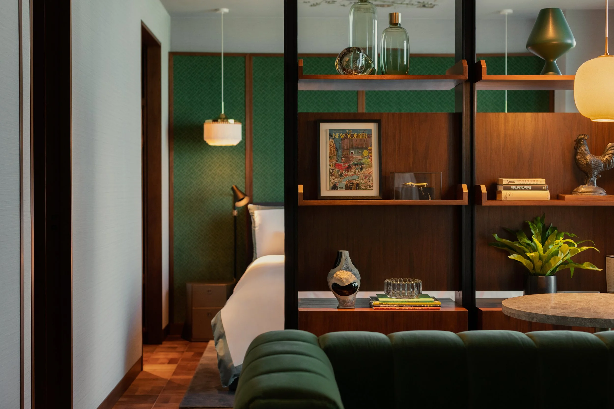Interior view of a modern living space, featuring a wooden room divider with decorative glass bottles, books, and sculptures, a green velvet chair, and hanging pendant lights with green and yellow shades.