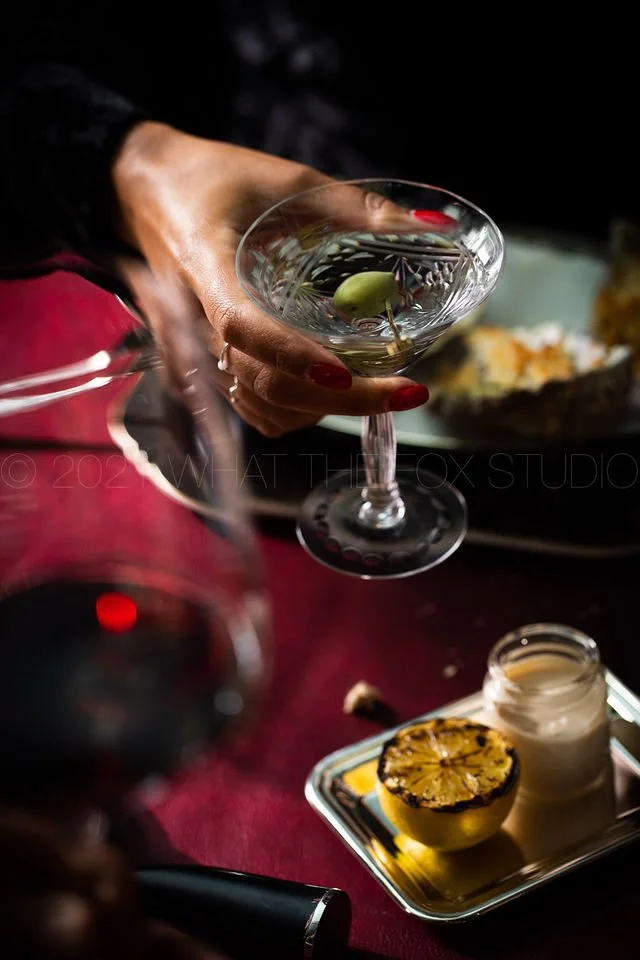 A woman with red painted nails holding a martini glass with an olive and cocktail pick. In the foreground, a blurred glass of red wine, and in the background, a plate with a grilled lemon half and a small container of white sauce on a tray. The table