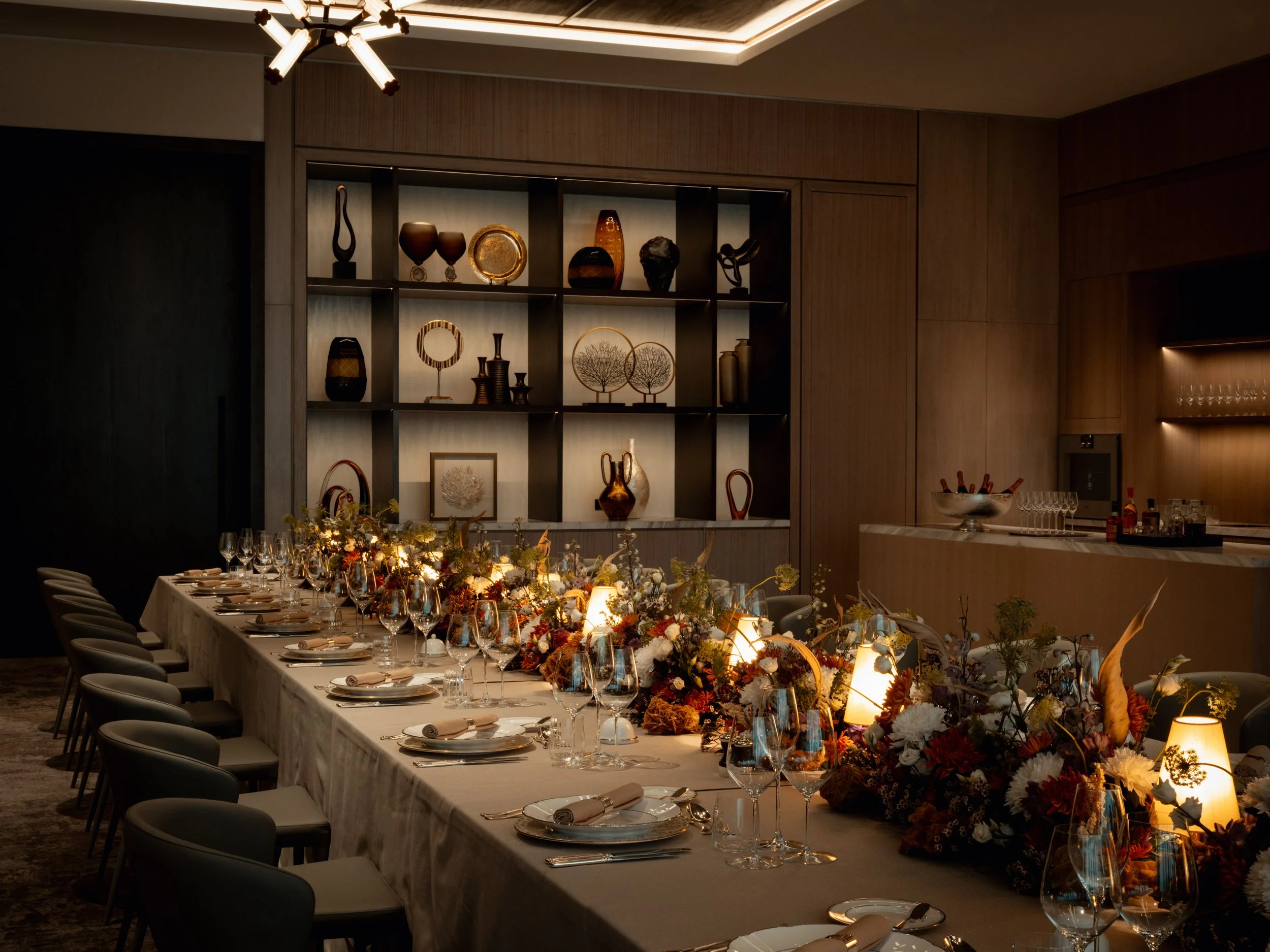A long banquet table decorated with a floral centerpiece, set with white plates, silverware, and wine glasses in a warmly lit, elegant dining room. The background features a wooden shelf with decorative pottery and sculptures, and a modern ceiling li