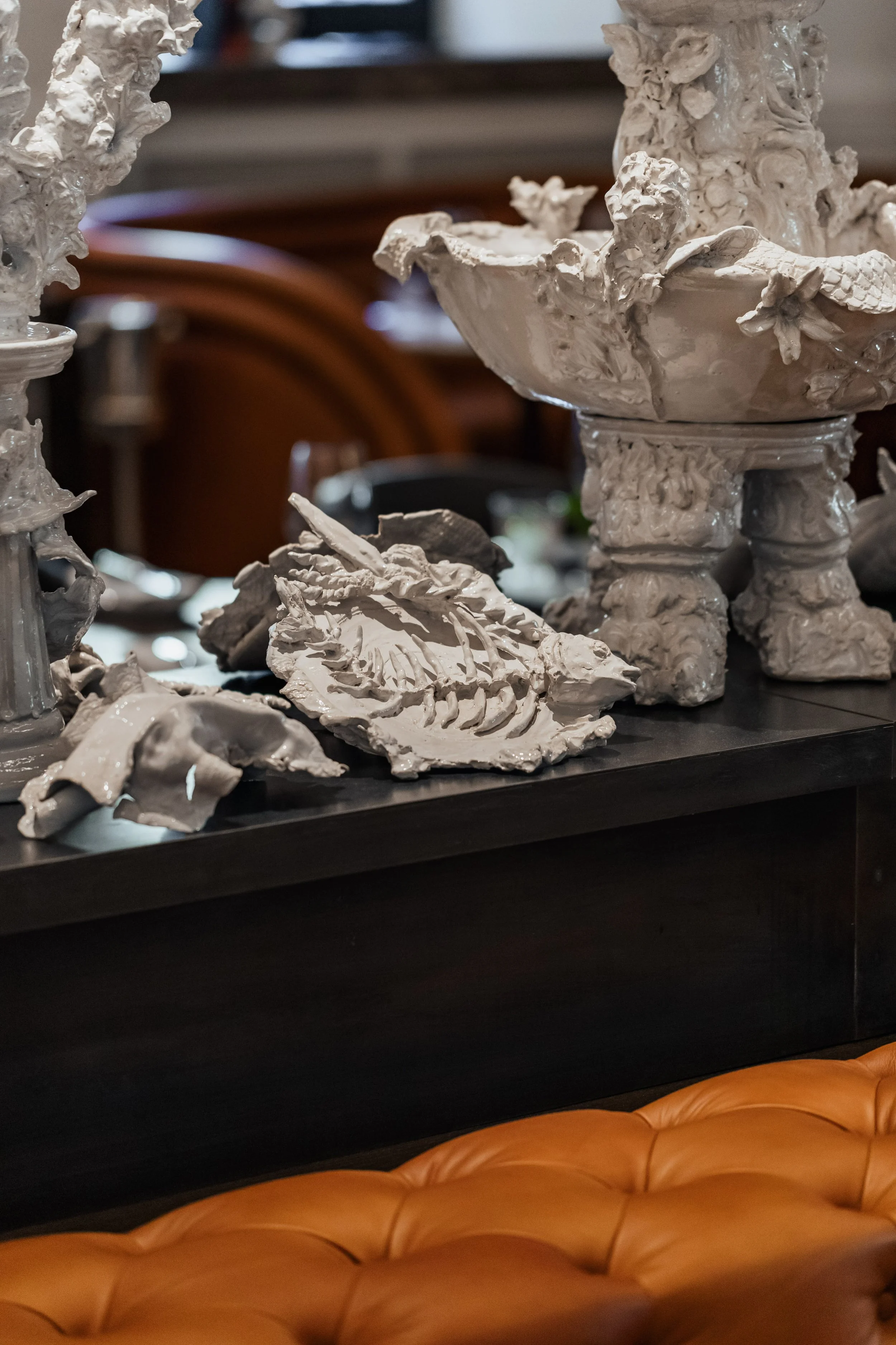 Close-up of white ceramic sculptures, including a large ornate bowl with flowers and a petal design, and a smaller sculpture resembling a leaf, placed on a dark table. In the background, there are wooden chairs and a brown leather couch at the bottom