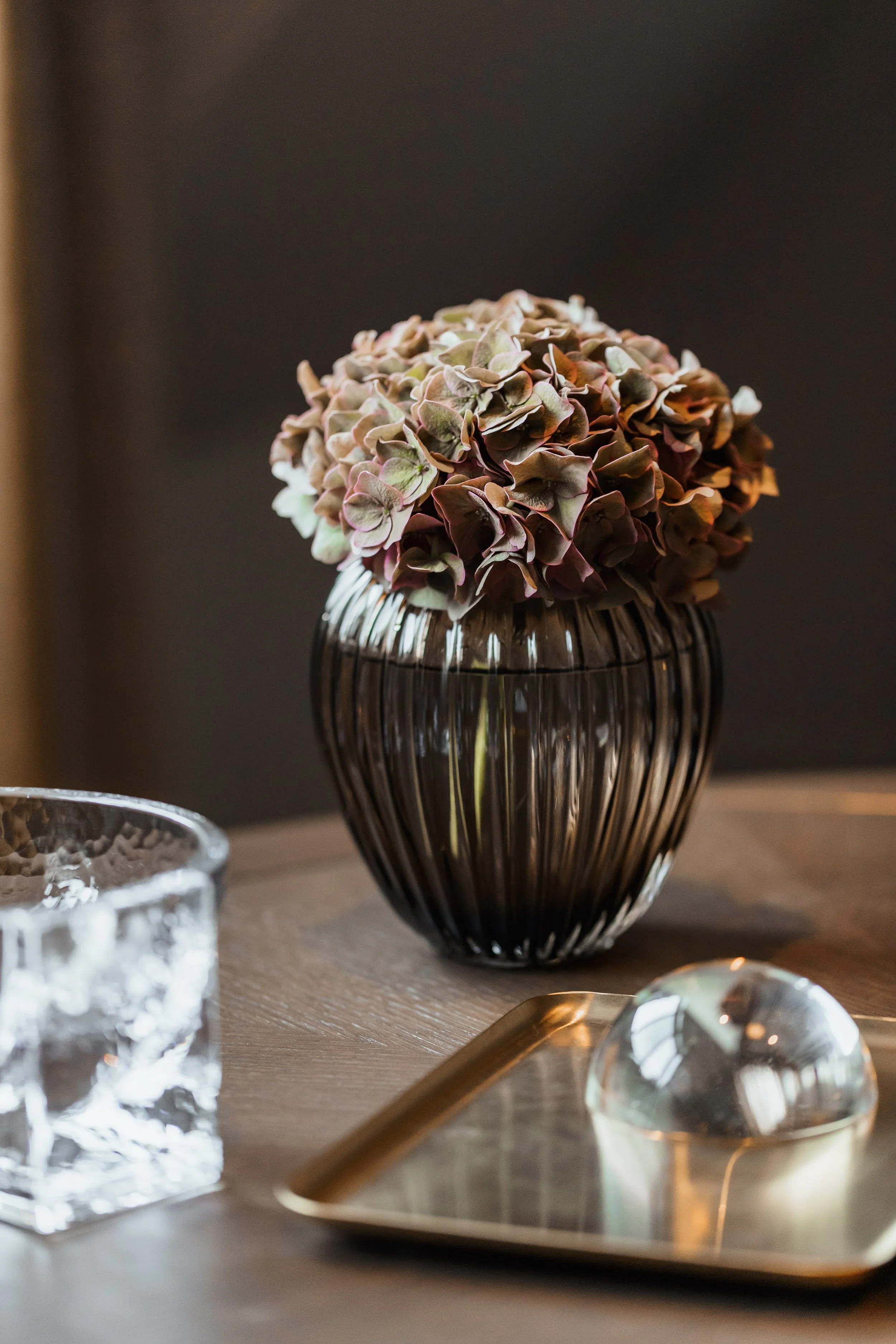 A black vase with a bouquet of pink and green hydrangeas on a wooden table, with a silver tray and glassware nearby.