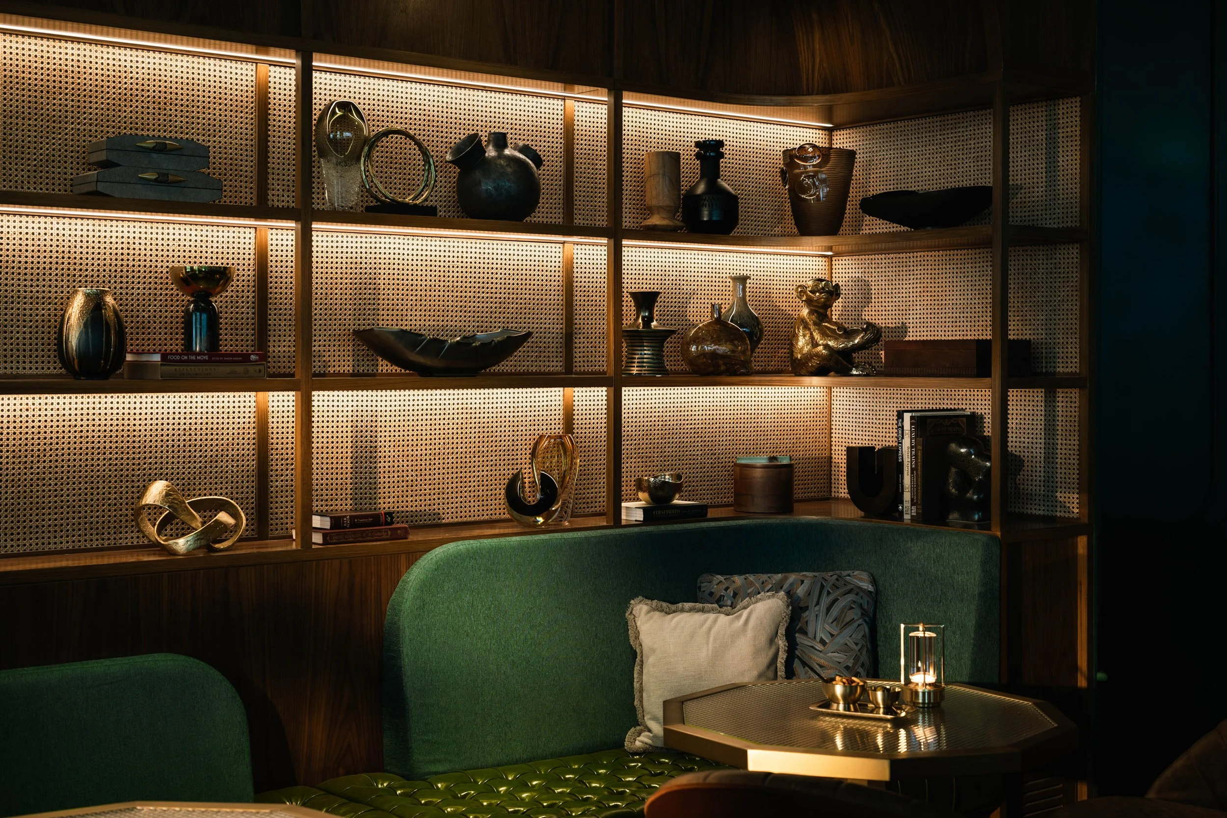 A living room with a wooden bookshelf displaying various decorative items, including vases, sculptures, and books, with green seating furniture in front.