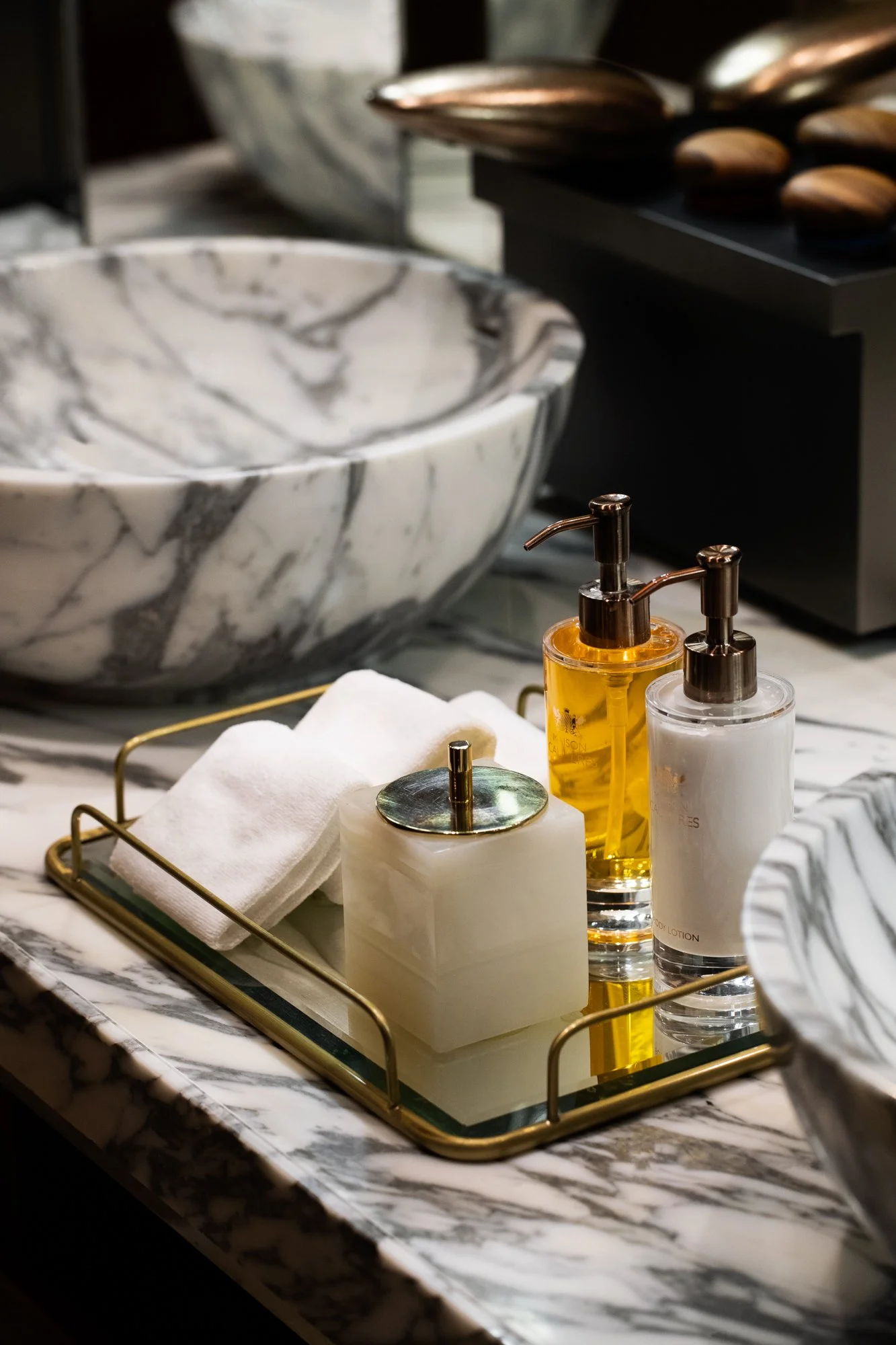 Bathroom countertop with a marble bowl sink, gold-colored soap and lotion dispensers, and a gold tray holding a white towel and a soap dish, all on a marble surface.