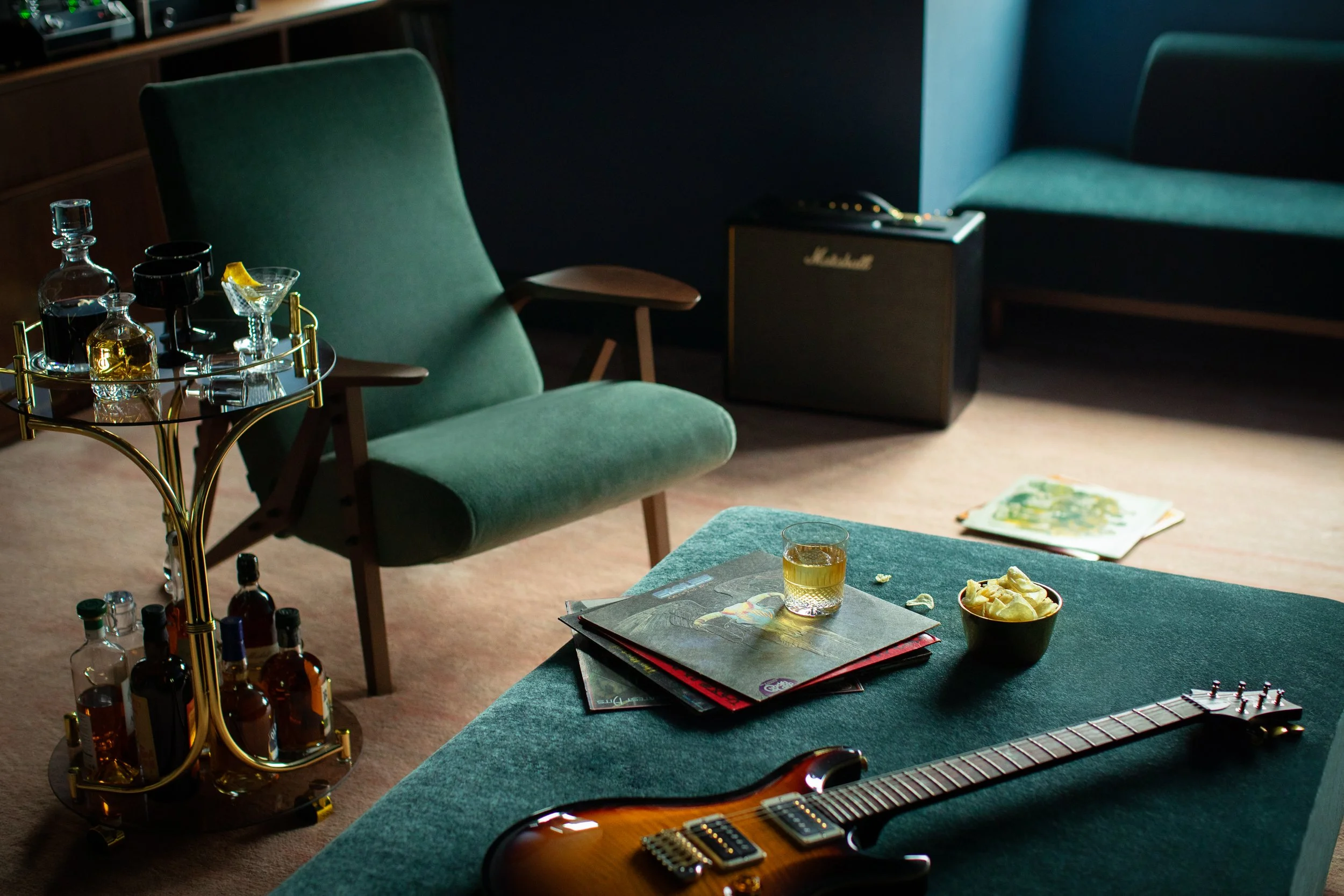 A cozy living room with green upholstered chairs, a small table with glassware and snacks, a guitar on the table, and a Marshall amplifier in the background. There are magazines or books on the table and a bowl of chips.