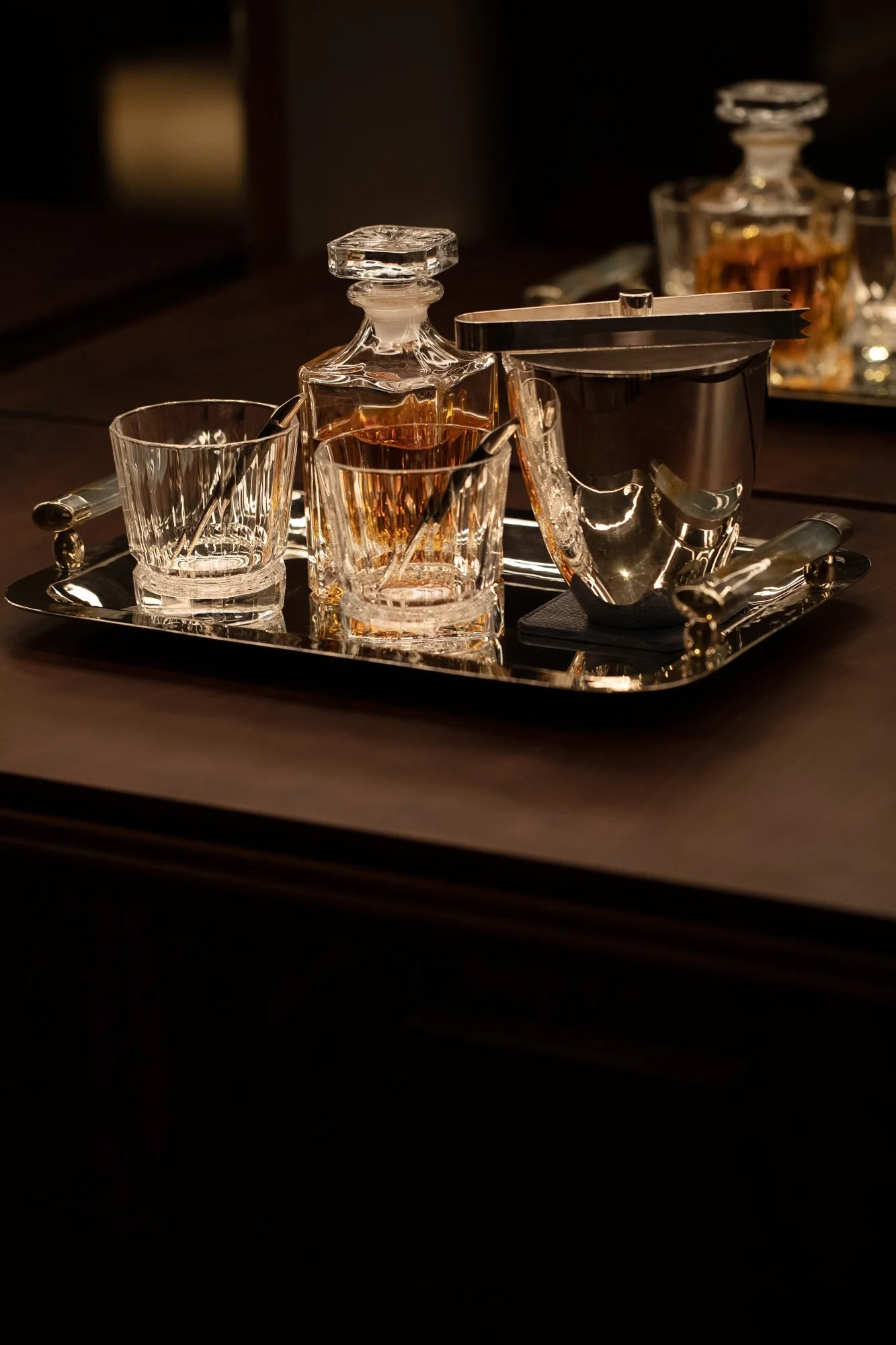 A tray with crystal decanters and glasses containing amber-colored liquids, likely whiskey, on a dark table with a blurred background.